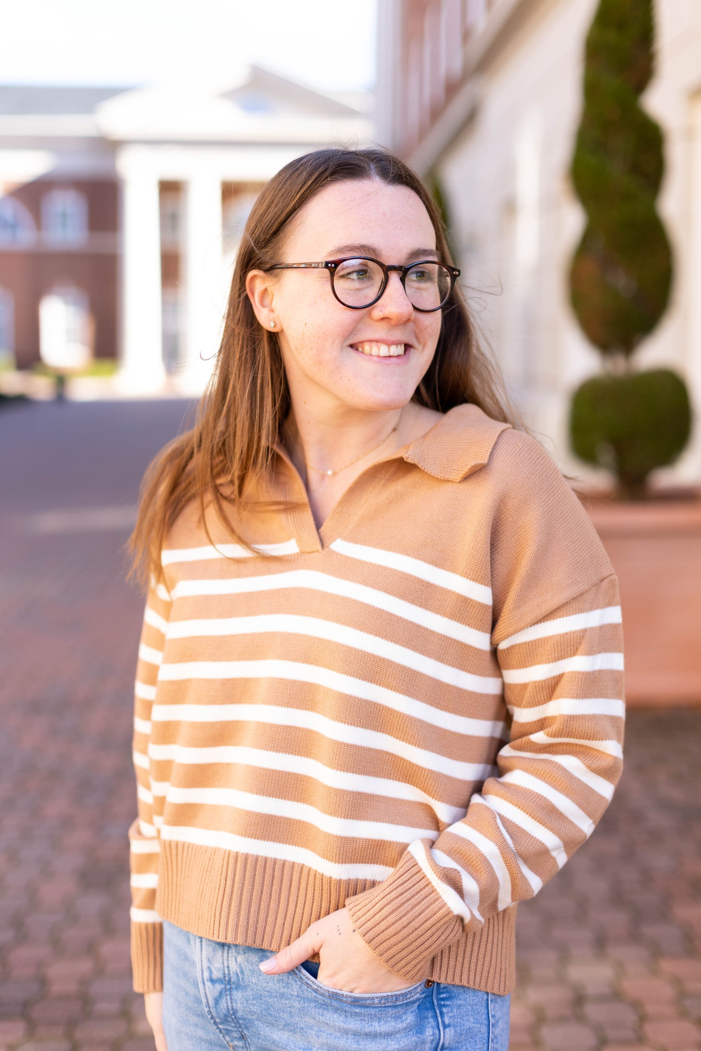 A person in glasses wears the chic Elliot Striped Sweater from Dogwood Cloth, featuring a brown and white design with a cropped fit. They stand outdoors, smiling with one hand in their pocket, as buildings and trees form the background.