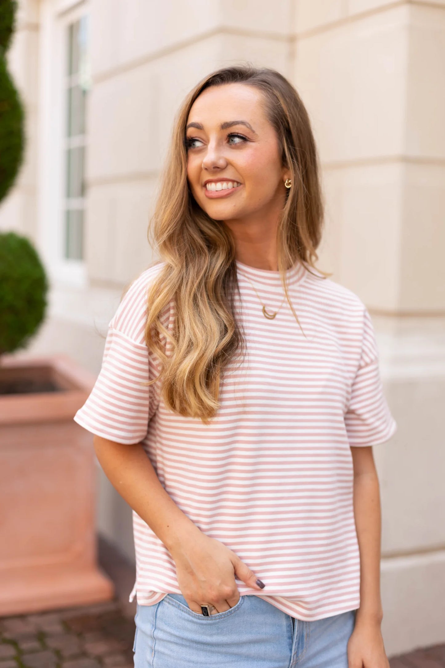 A woman with long hair smiles, wearing a Dogwood Cloth Delaney Striped Tee and jeans, exuding casual comfort as she stands in front of a building with plants in the background.