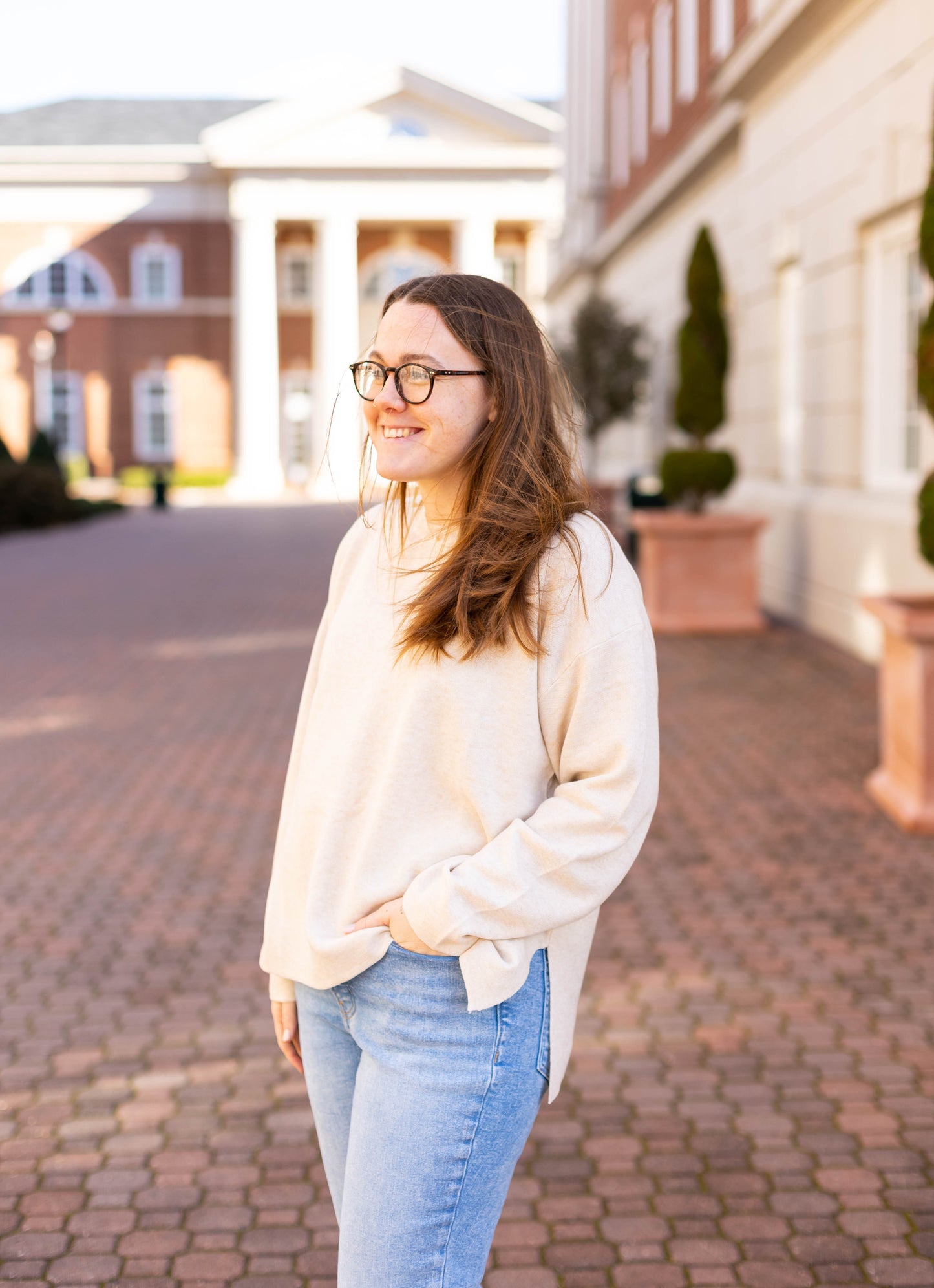 Standing outdoors on a brick pathway, a person with long hair and glasses exudes effortless elegance in a beige Winslet Long-Sleeve sweater from Dogwood Cloth, crafted from luxe-feel fabric, paired with jeans. In the background, buildings rise into the clear sky.