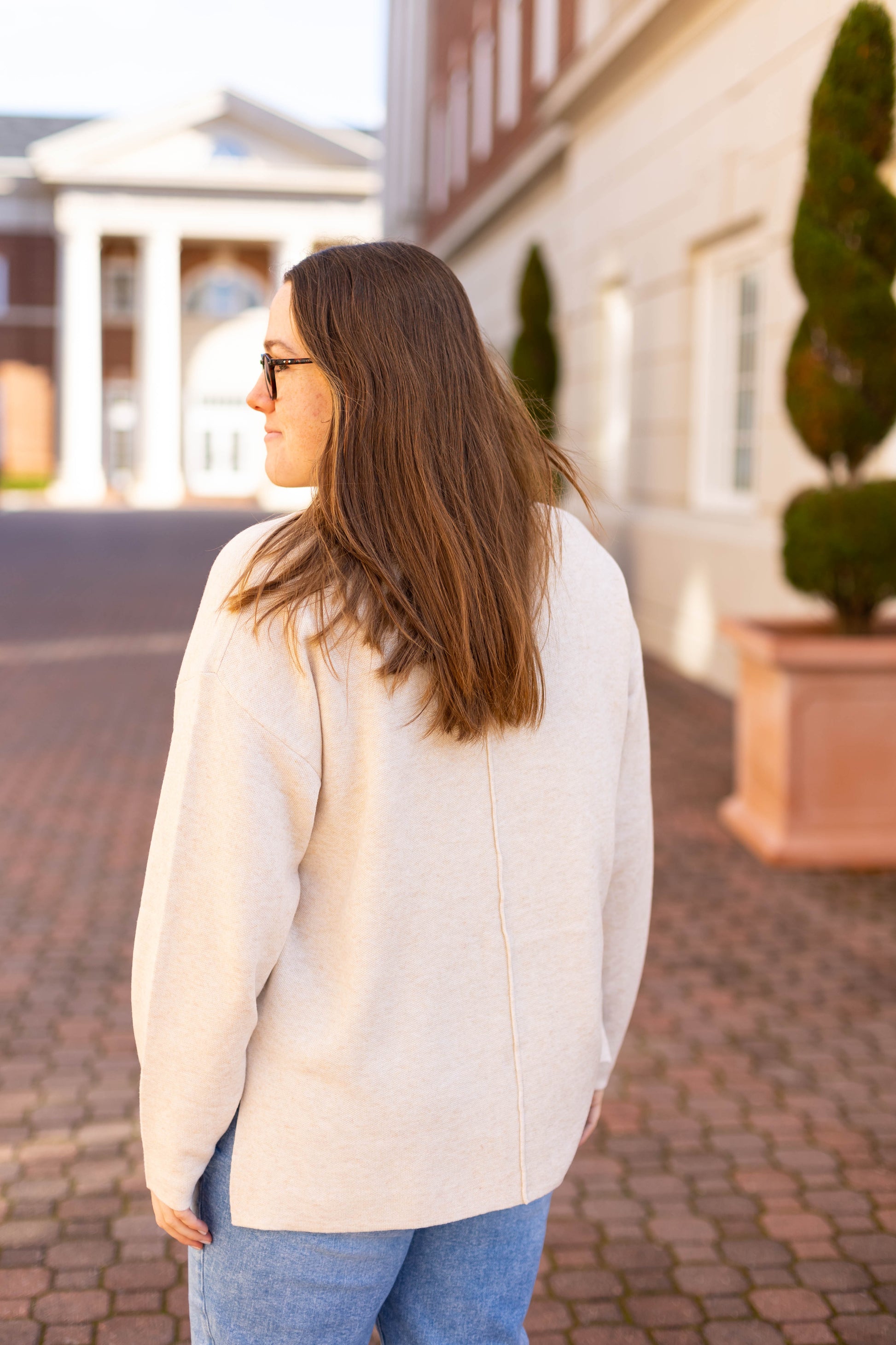 A person with long brown hair, dressed in a beige Dogwood Cloth Winslet Long-Sleeve and jeans, stands gracefully outdoors near a columned building.