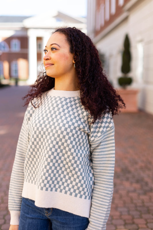 A person with curly hair, dressed in the Emory Contrast Sweater by Dogwood Cloth, stands outdoors on a brick pathway, gazing to the side.