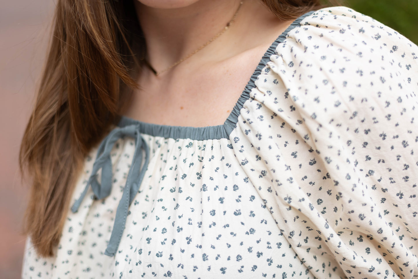 Close-up of someone wearing the Dogwood Cloth Kate Floral Top, a romantic blouse with a white base, blue floral print, and a blue-trimmed square neckline.