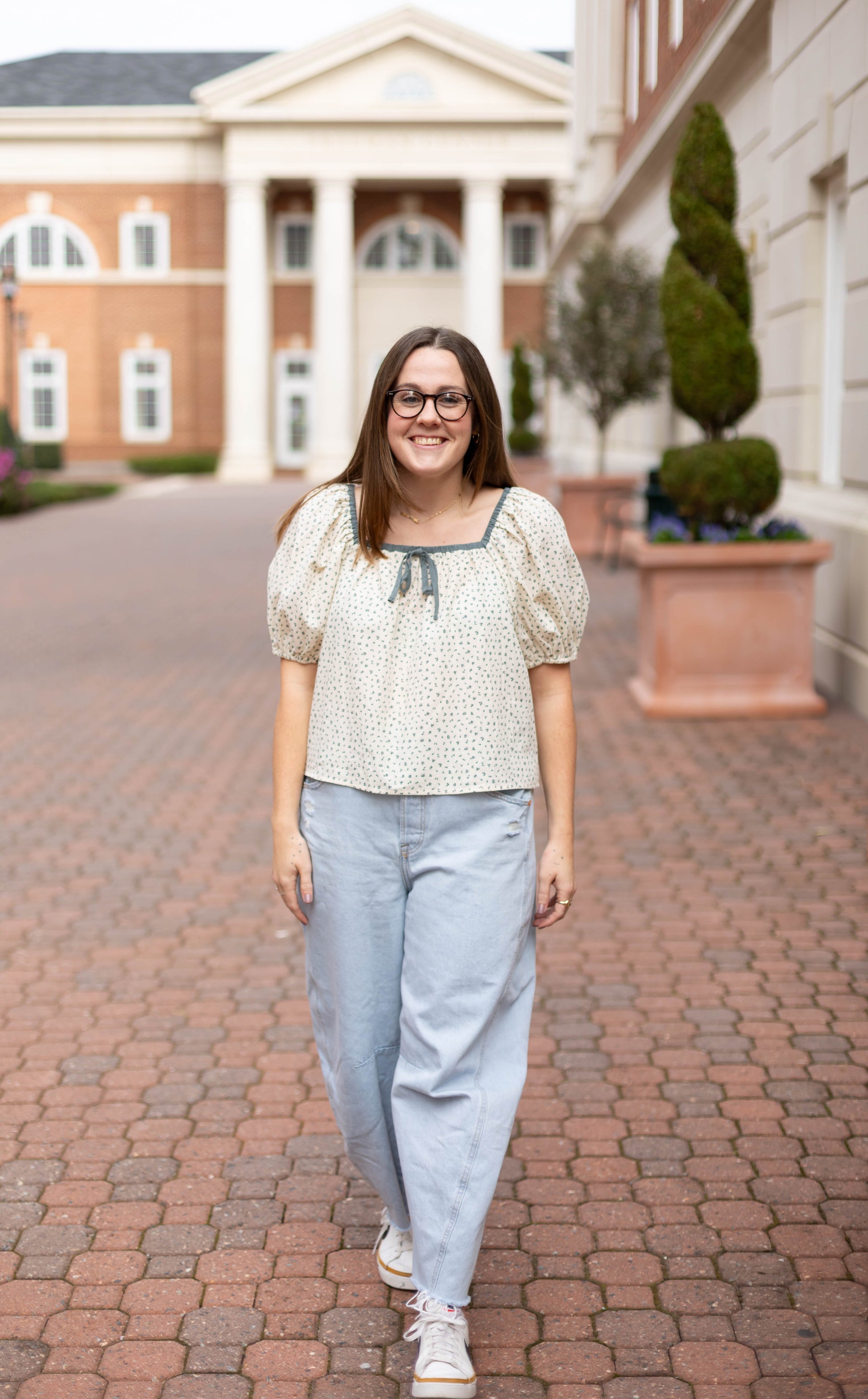 A woman with long brown hair and glasses, wearing the Dogwood Cloth Kate Floral Top and light blue jeans, walks on a brick pathway between two buildings.