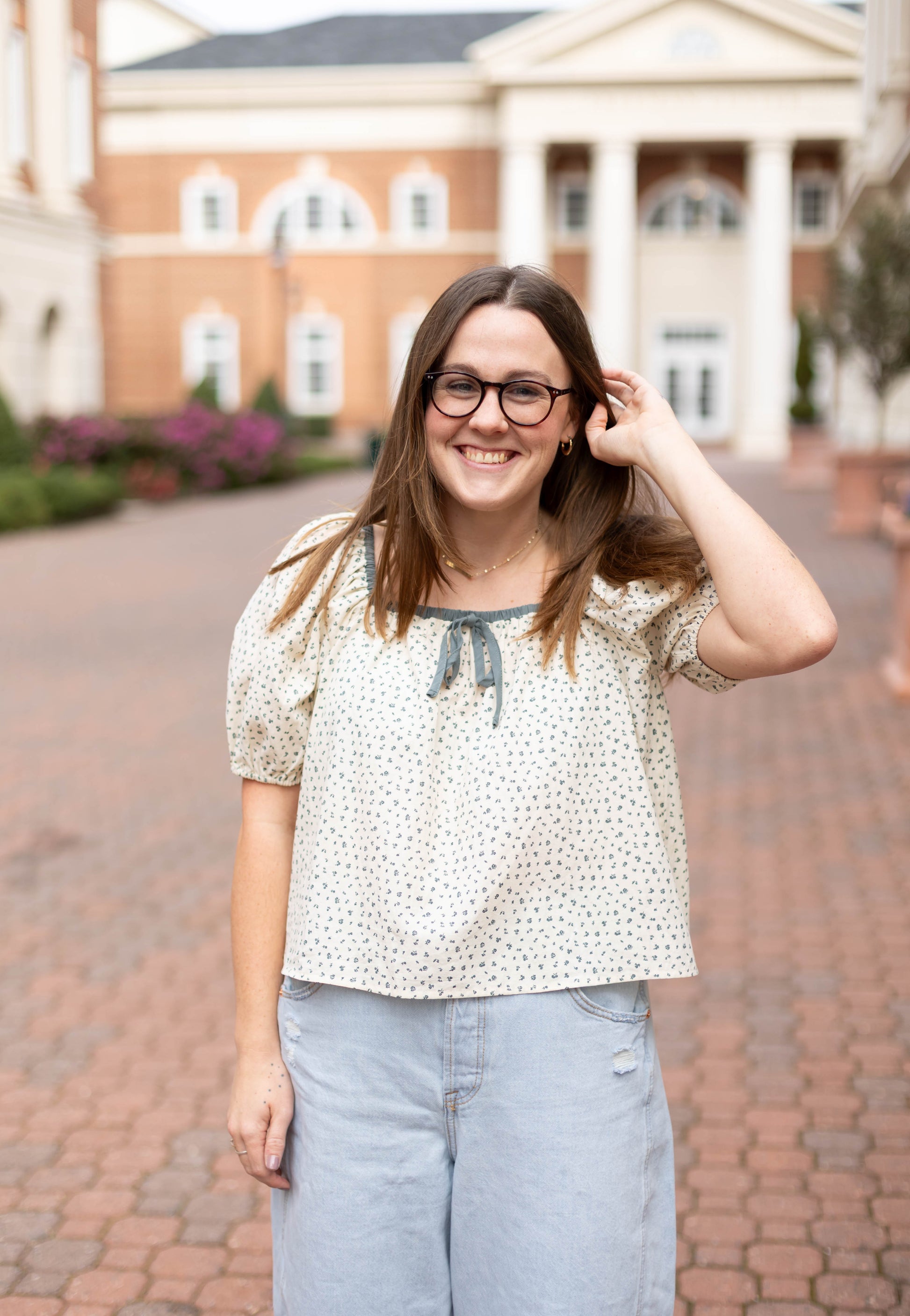 A woman with long brown hair, glasses, a Dogwood Cloth Kate Floral Top, and light jeans stands smiling outdoors in front of a brick building with white columns.