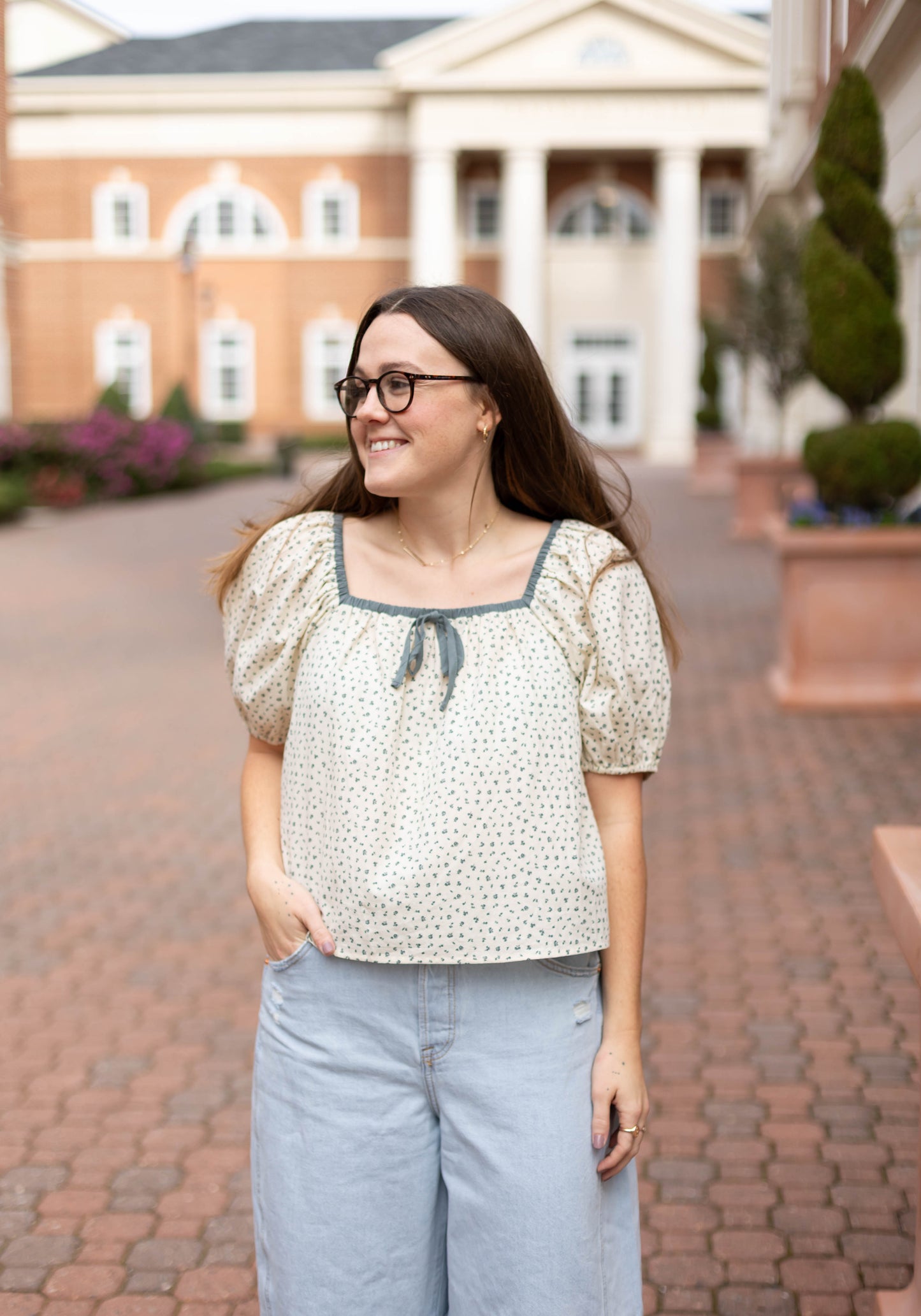 A woman in glasses and light blue jeans wears the Dogwood Cloth Kate Floral Top while standing outdoors on a brick walkway in front of a building with columns.