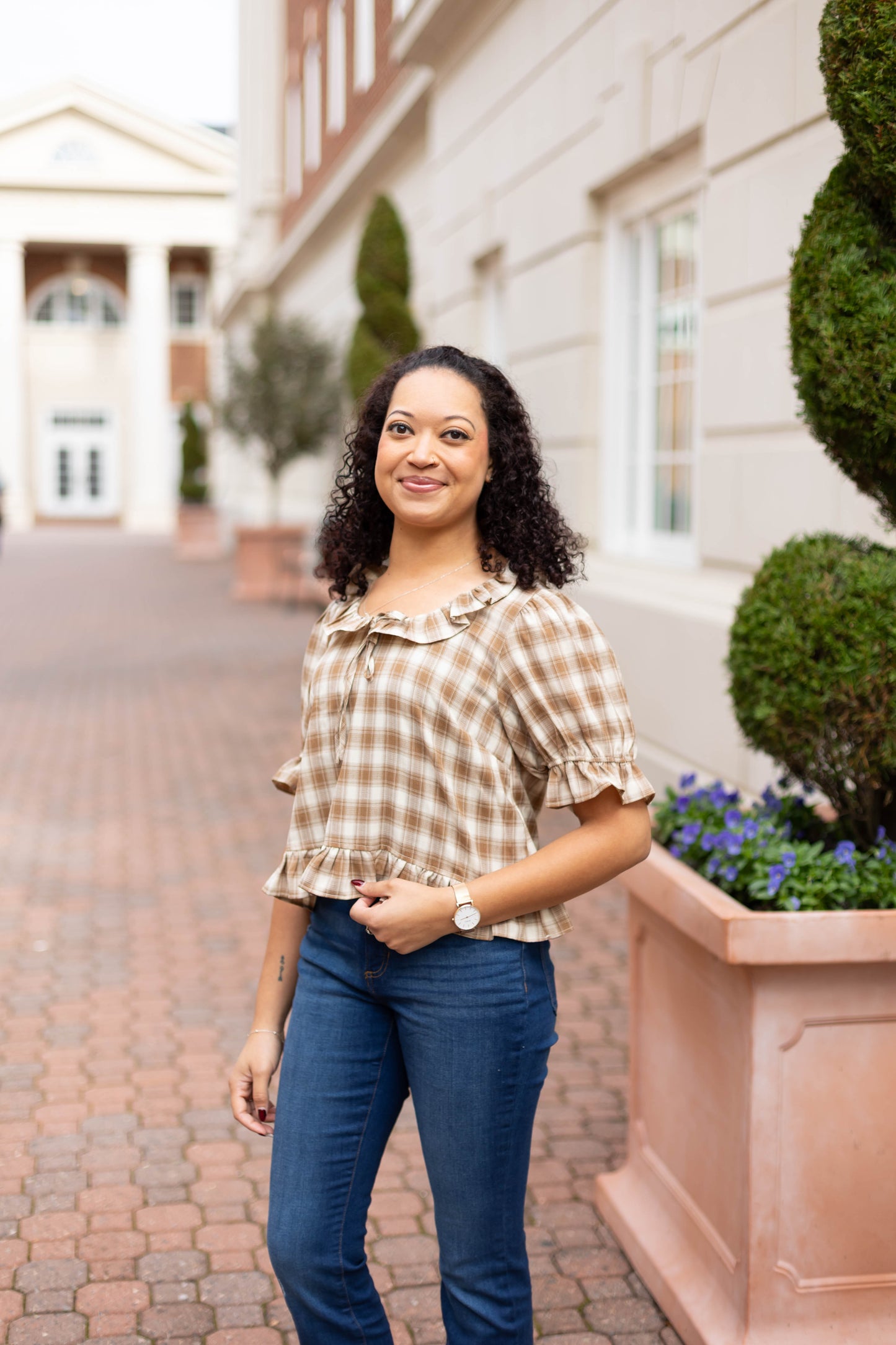 A woman with curly hair wearing the Addie Plaid Blouse by Dogwood Cloth and blue jeans stands on a brick walkway beside a white building with large planters.
