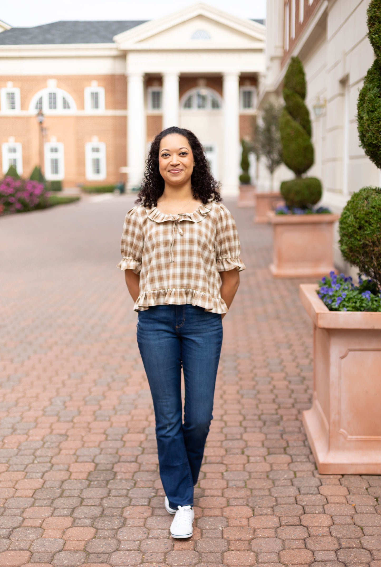 A woman with curly hair stands outdoors on a brick walkway, hands in pockets, wearing the Dogwood Cloth Addie Plaid Blouse with blue jeans and white shoes. Buildings and planters appear in the background.