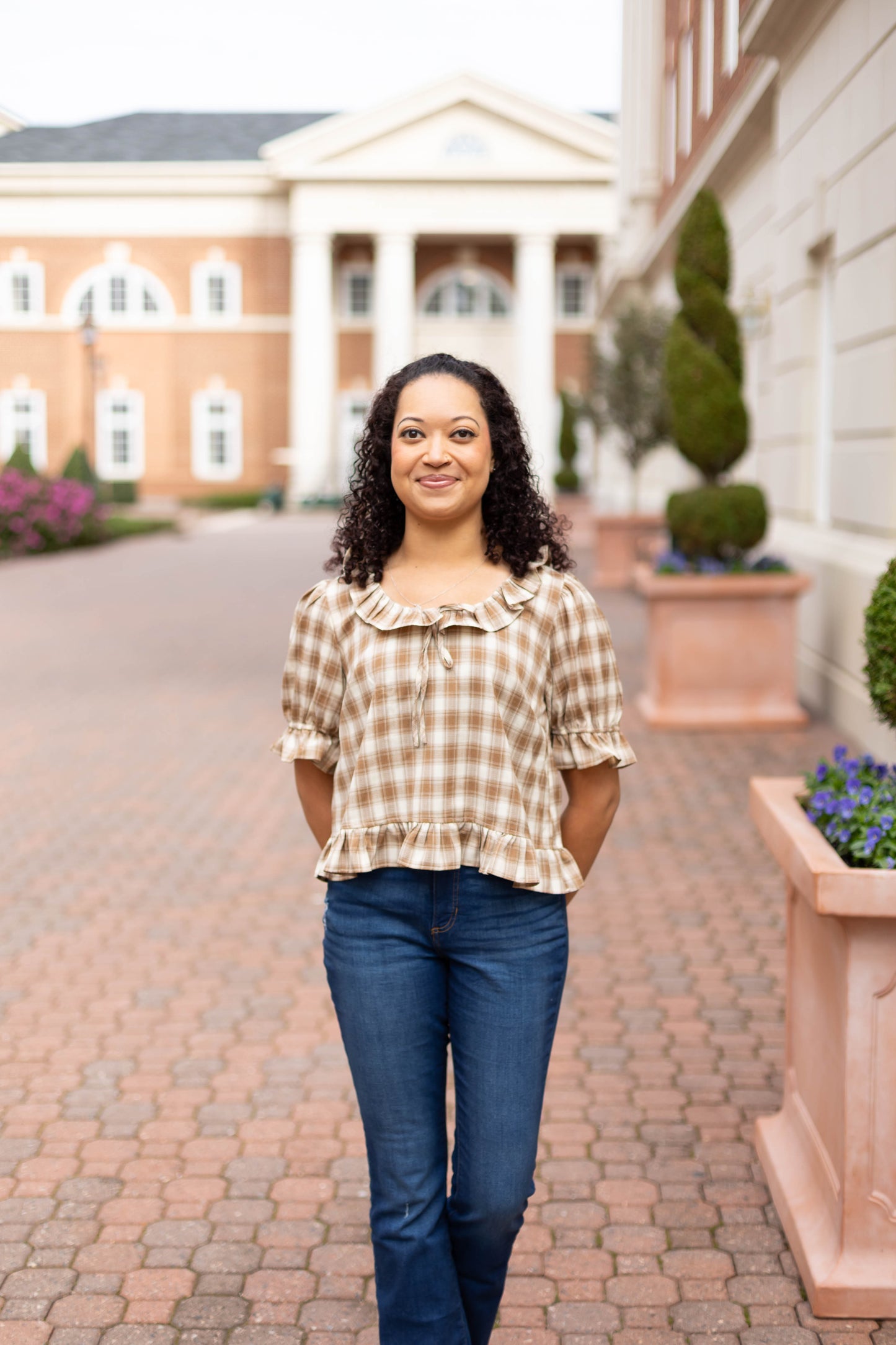 A woman with curly hair stands on a brick walkway outside a columned building, wearing the Addie Plaid Blouse by Dogwood Cloth and jeans, hands behind her back.
