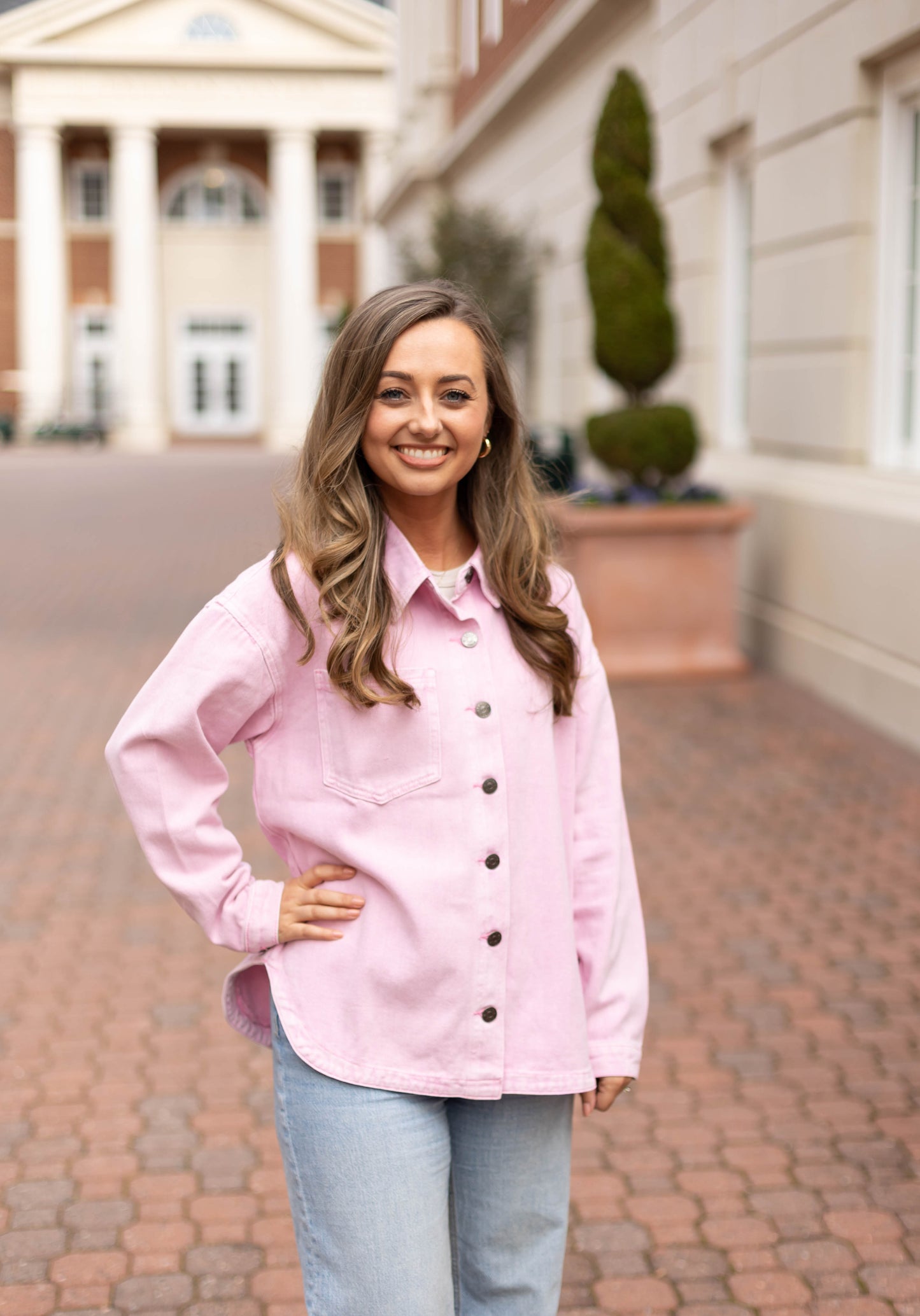 A woman with long brown hair smiles at the camera while standing outdoors on a brick walkway, wearing the Leah Denim Jacket by Dogwood Cloth—an essential addition to any wardrobe.