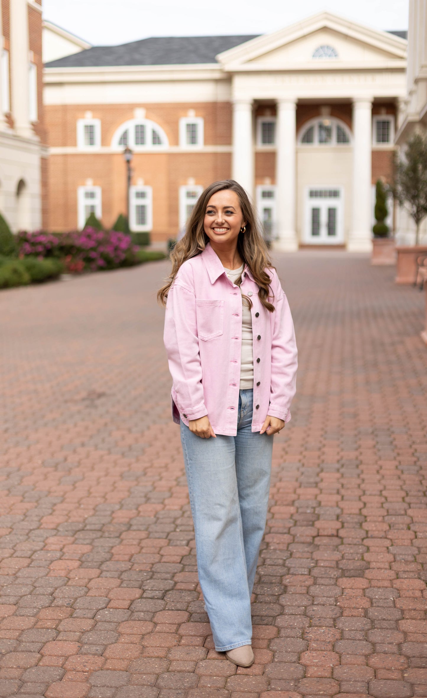 A woman stands on a brick walkway in front of a building with columns, wearing the Dogwood Cloth Leah Denim Jacket over a striped shirt, wide-leg jeans, and beige shoes.