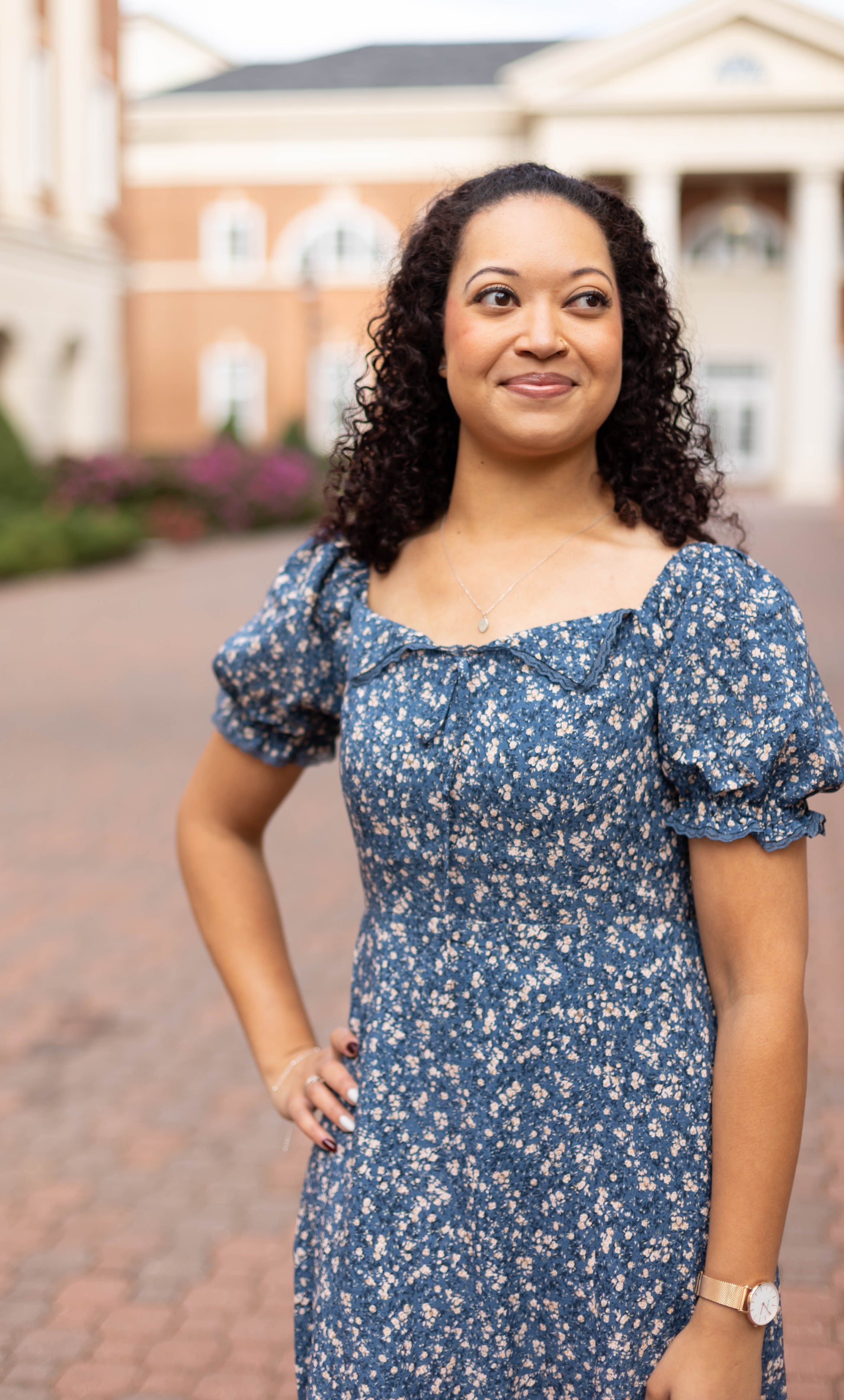 A woman stands outdoors on a paved walkway, framed by buildings and greenery, wearing the Olivia Floral Midi dress by Dogwood Cloth.