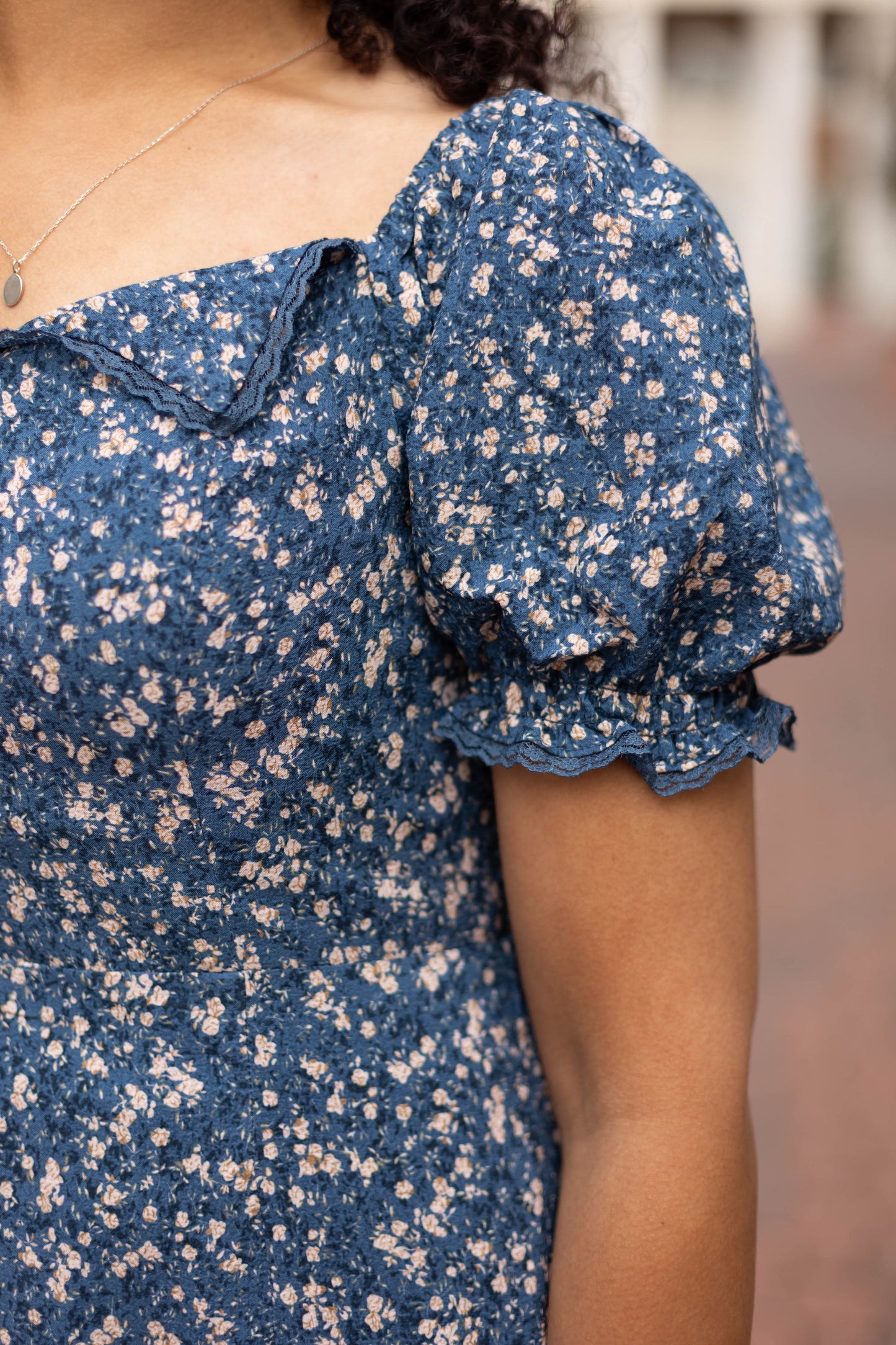 A person wearing the Dogwood Cloth Olivia Floral Midi dress, featuring puffed sleeves and a smocked back, paired with a pendant necklace, shown from the shoulders down.