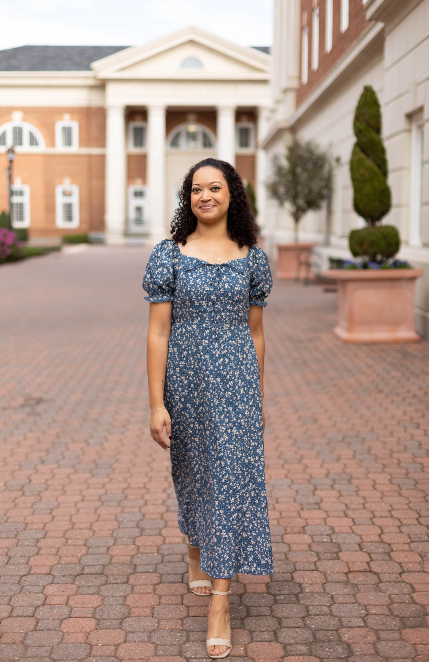 A woman in the Olivia Floral Midi by Dogwood Cloth, a blue dress with a lace-trimmed collar, walks along a brick pathway between columned buildings and manicured shrubs.