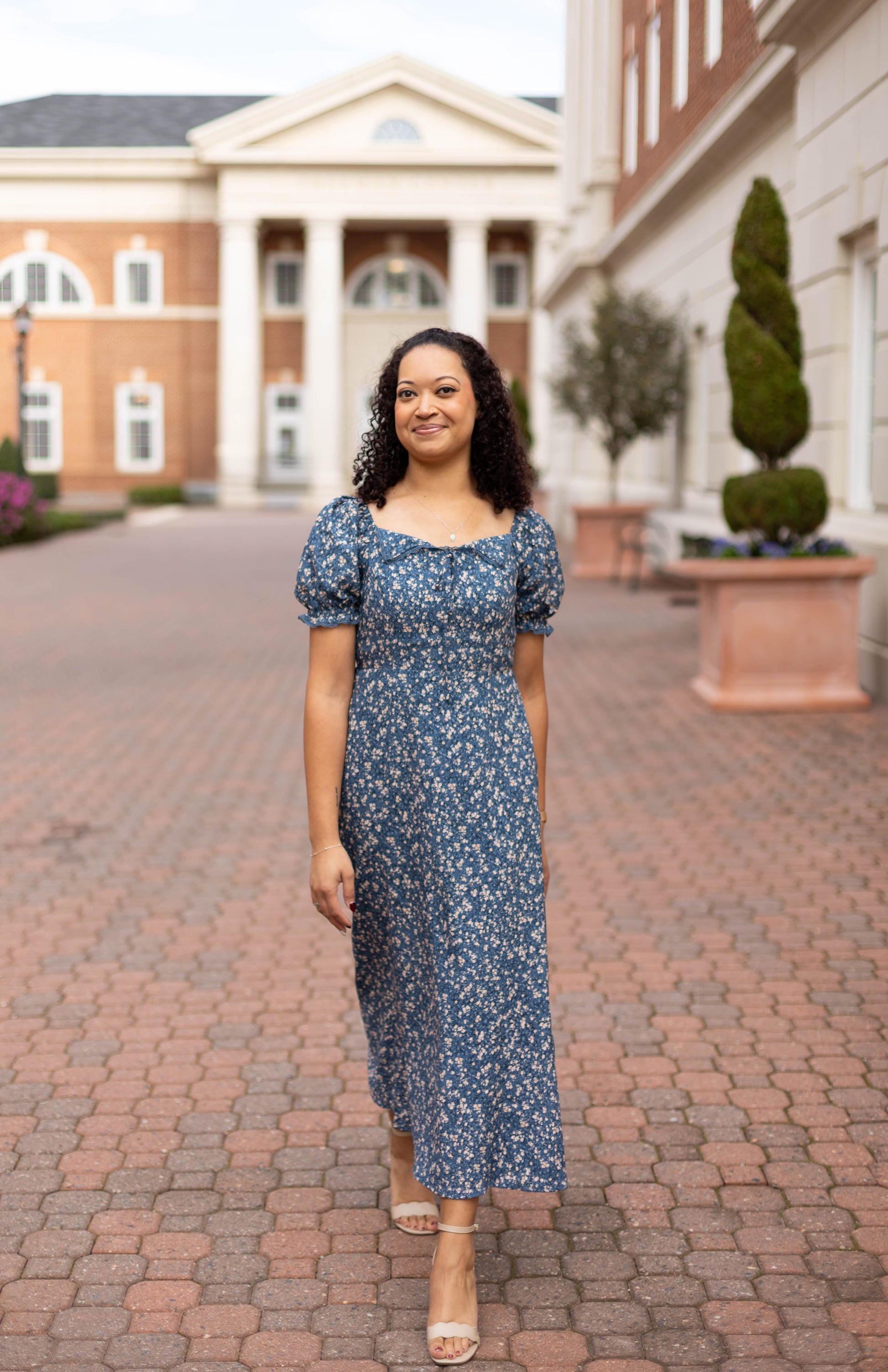 A woman in the Olivia Floral Midi by Dogwood Cloth, a blue dress with a lace-trimmed collar, walks along a brick pathway between columned buildings and manicured shrubs.
