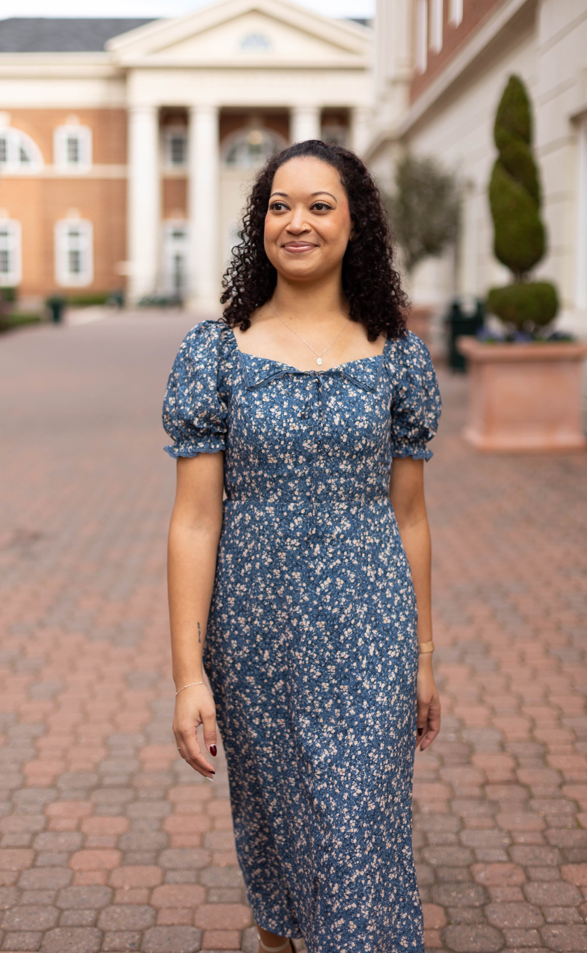 A woman with curly hair wears the Olivia Floral Midi by Dogwood Cloth as she walks on a brick pathway in front of a building with white columns.