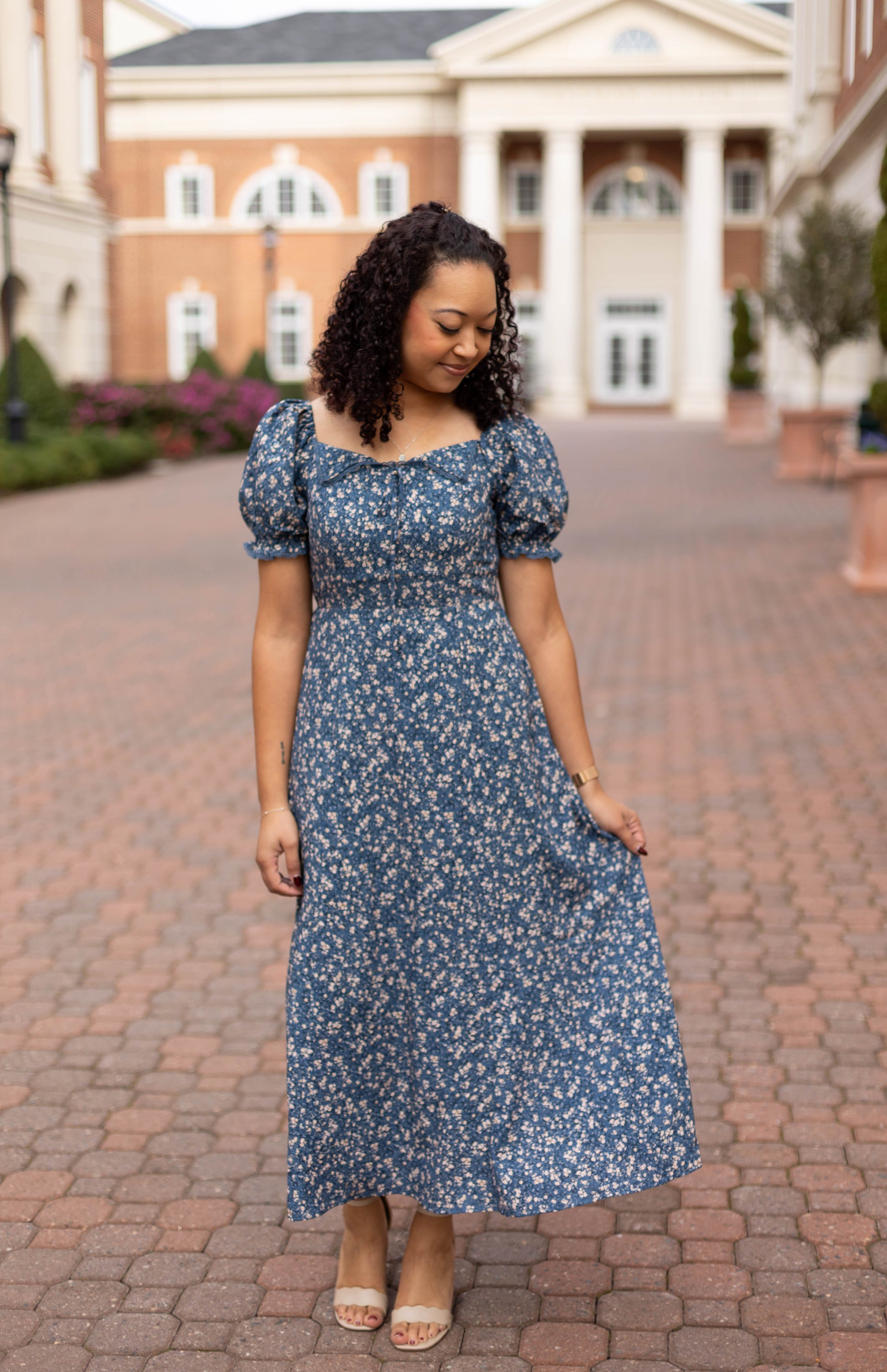 A woman with curly hair wears the Olivia Floral Midi by Dogwood Cloth, featuring a blue floral print and lace-trimmed collar, as she stands on a brick walkway gently holding her dress and looking down.