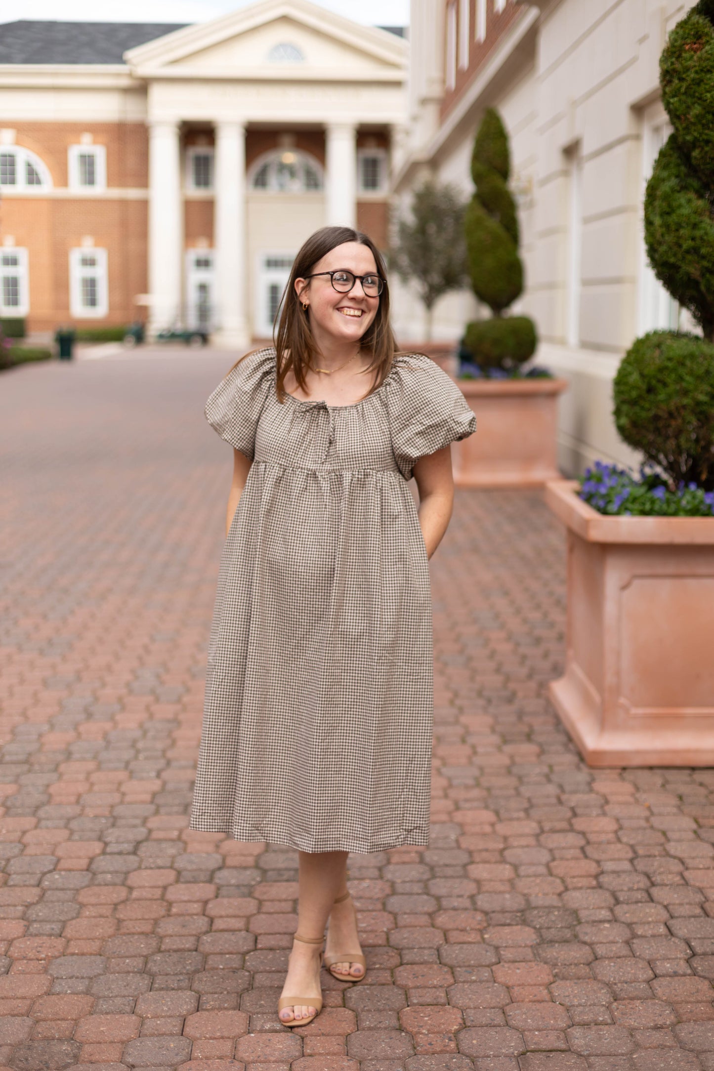 A woman in glasses and a light brown Dogwood Cloth Nicole Gingham Midi smiles on a brick walkway, with large potted plants and a columned building in the background.