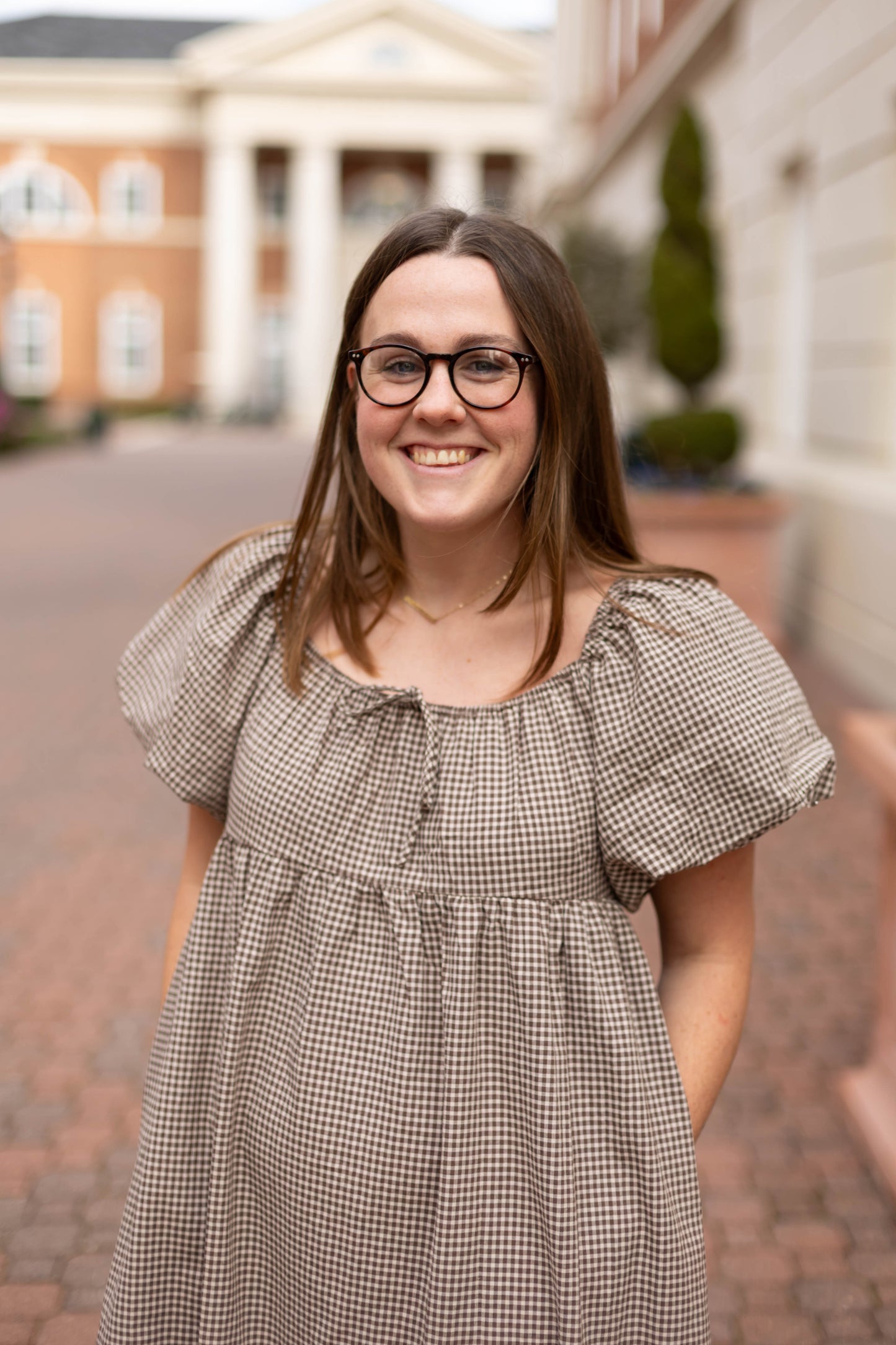 A woman with long brown hair and glasses smiles outdoors on a brick path, wearing the Nicole Gingham Midi by Dogwood Cloth. A building with columns is visible in the background.