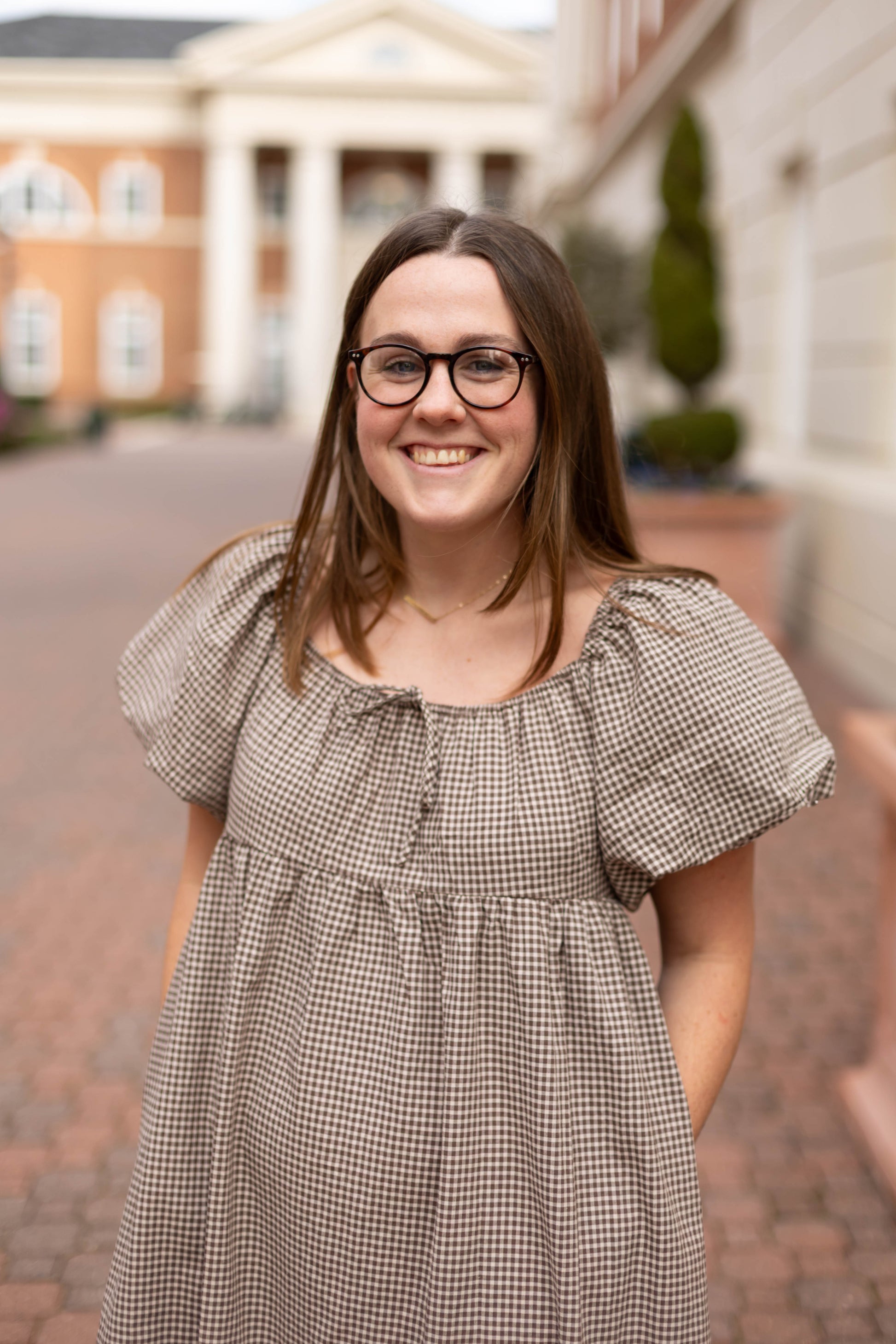 A woman with long brown hair and glasses smiles outdoors on a brick path, wearing the Nicole Gingham Midi by Dogwood Cloth. A building with columns is visible in the background.