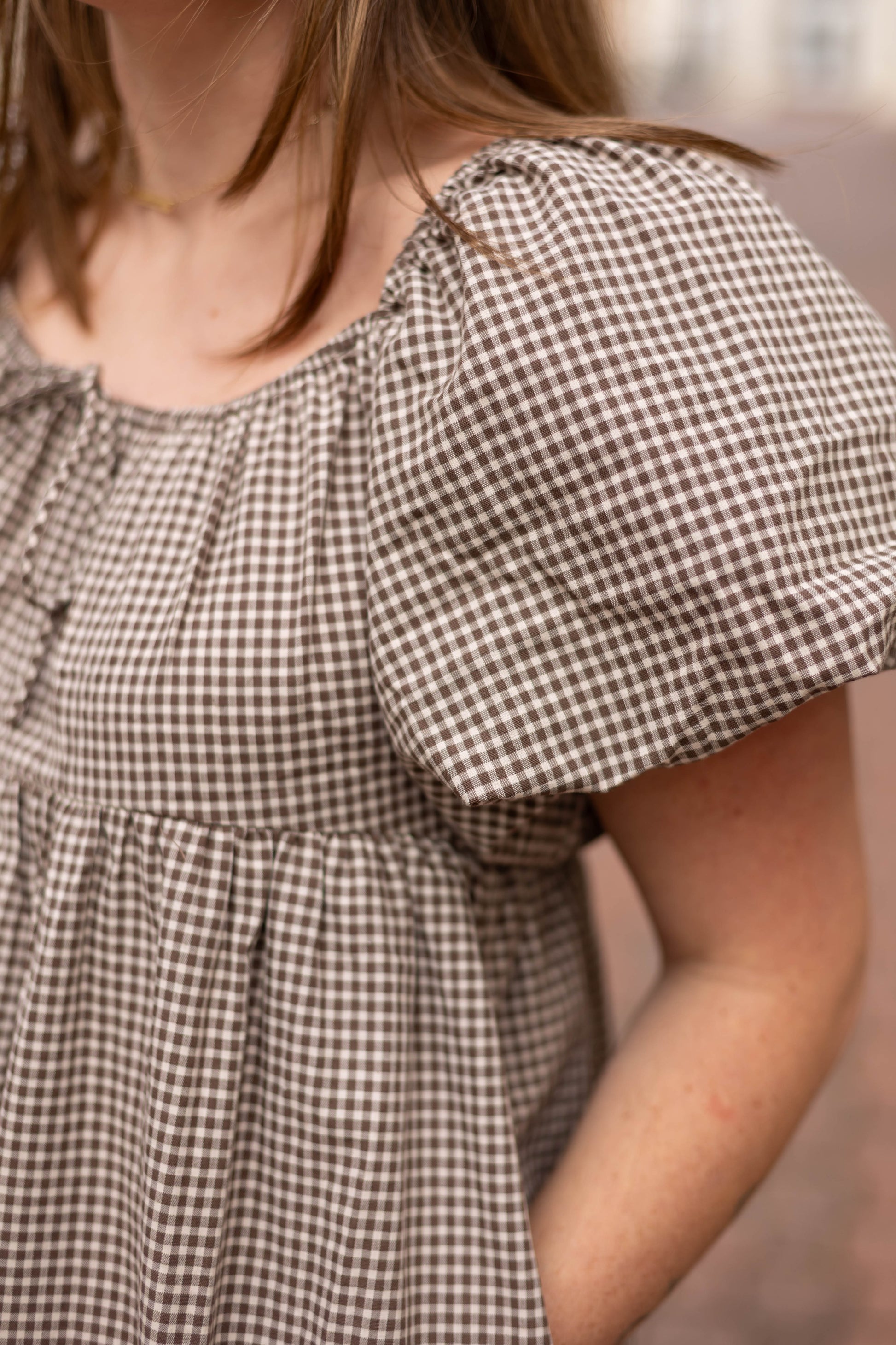 Someone stands outdoors with a hand in their pocket, wearing the Nicole Gingham Midi dress by Dogwood Cloth; only the upper body and part of one arm are visible.