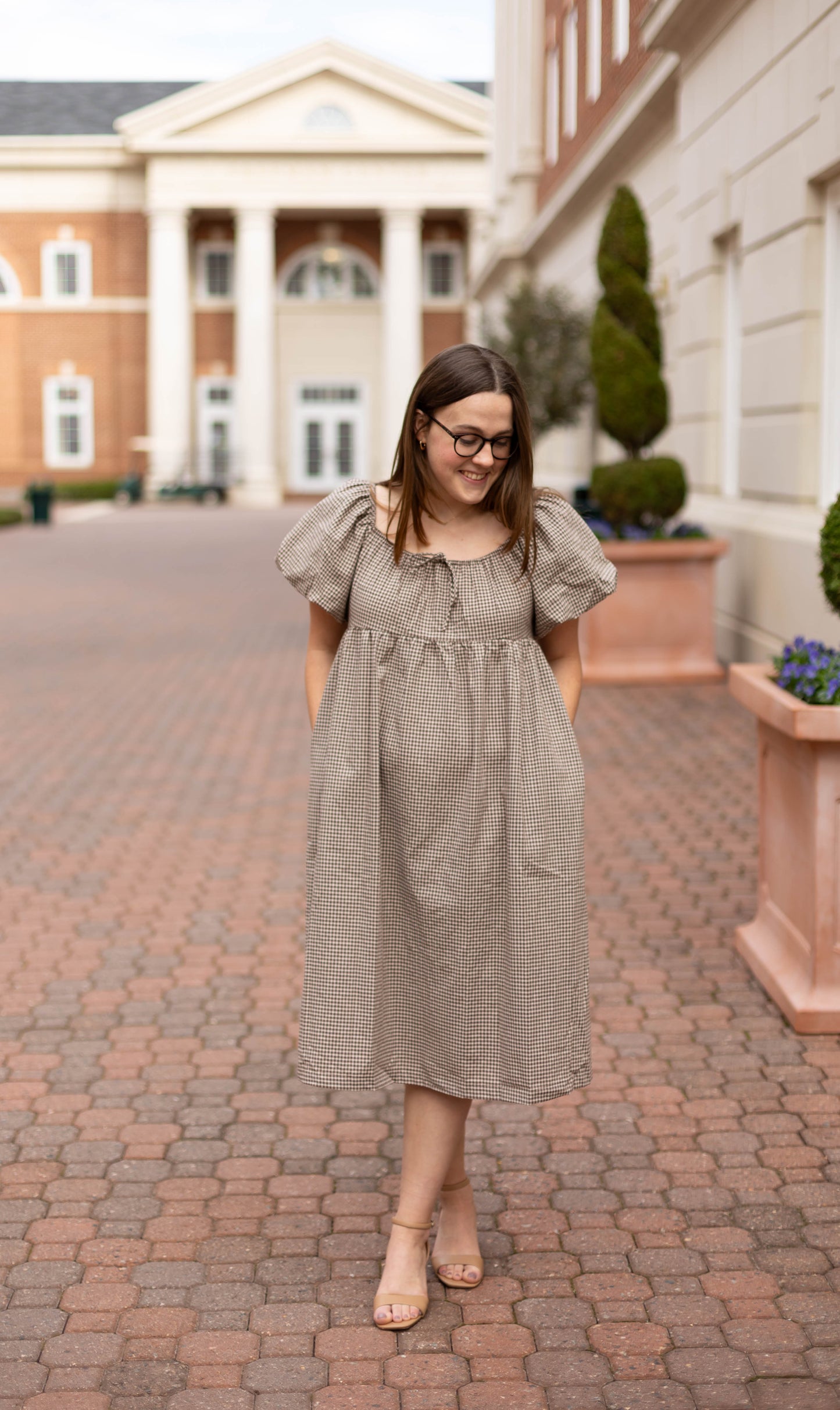 A woman wears the Nicole Gingham Midi by Dogwood Cloth, standing with hands in pockets on a brick walkway before a columned building.