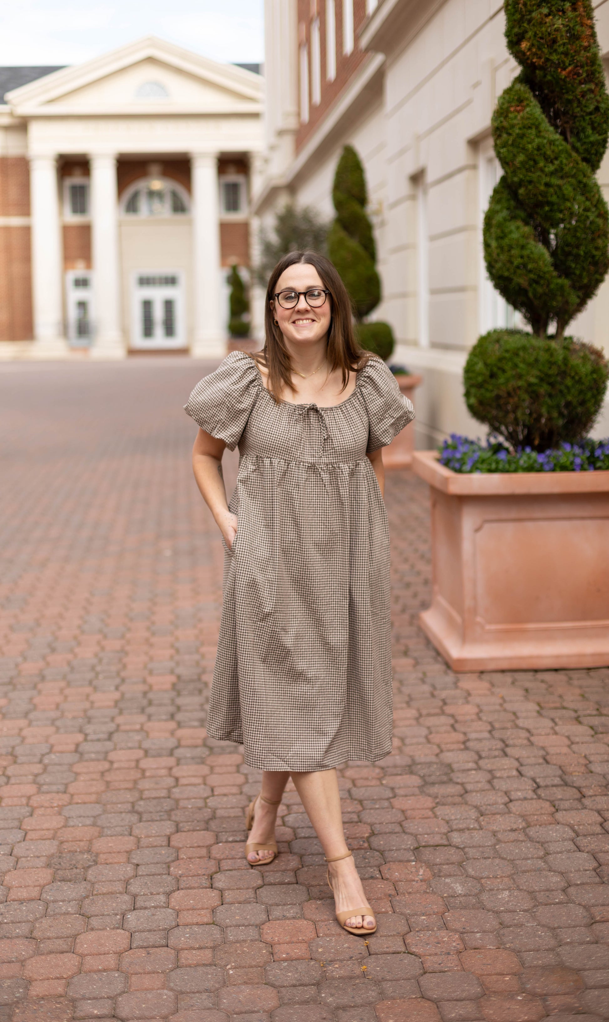 A woman in the Nicole Gingham Midi by Dogwood Cloth and sandals stands on a brick walkway outside a building with columns and manicured bushes.