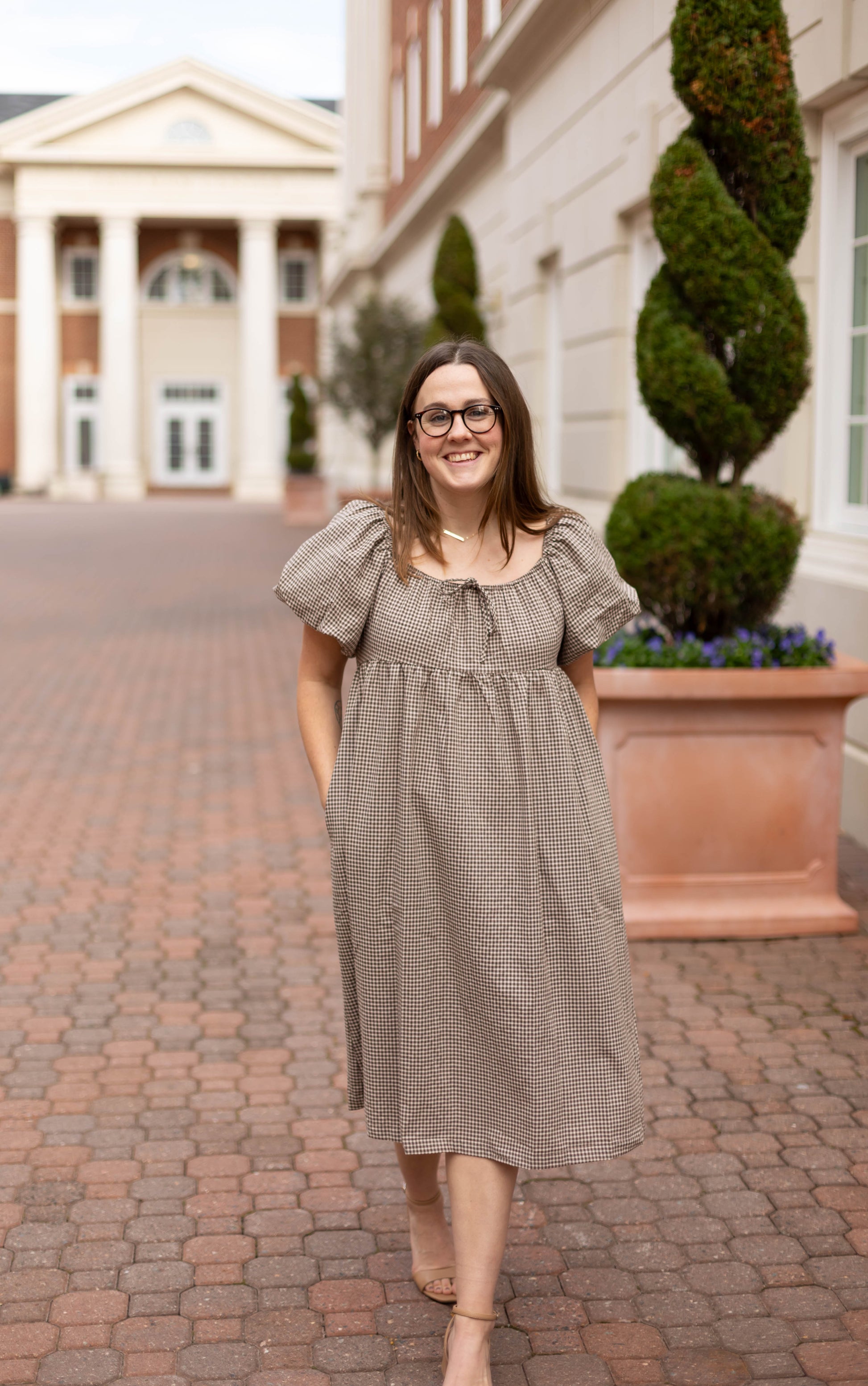 A woman wears the Dogwood Cloth Nicole Gingham Midi, featuring puff sleeves, as she smiles at the camera while walking on a brick pathway beside a columned building and manicured shrubs.