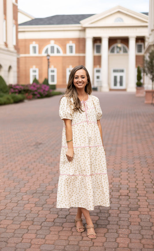 A woman wears the Dogwood Cloth Abigail Floral Midi, smiling on a brick pathway in front of a white-columned building.