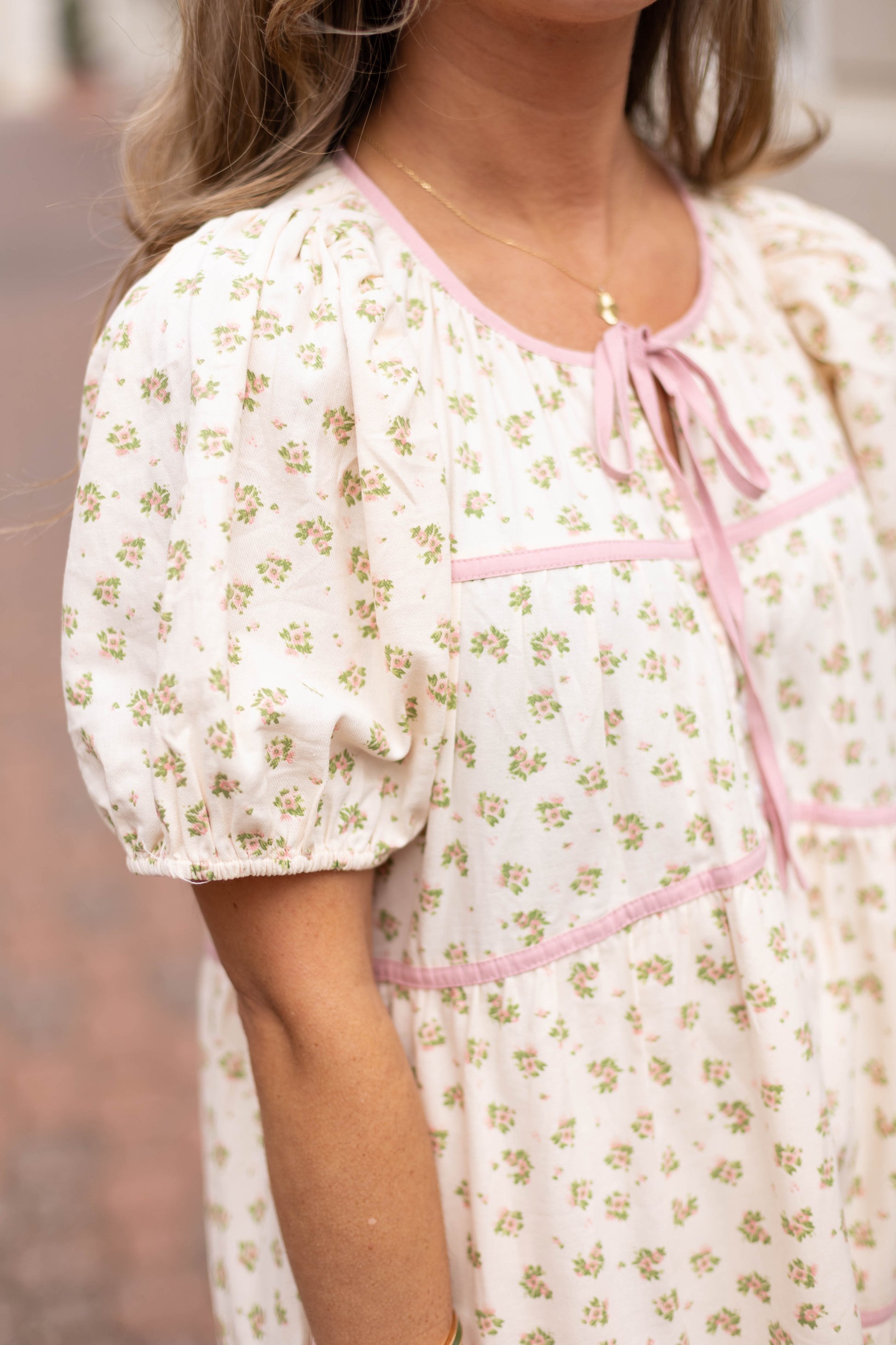 The Abigail Floral Midi by Dogwood Cloth features white cotton with green and pink florals, pink trim, puffed sleeves, and a neckline bow—perfect for a charming cottagecore look.