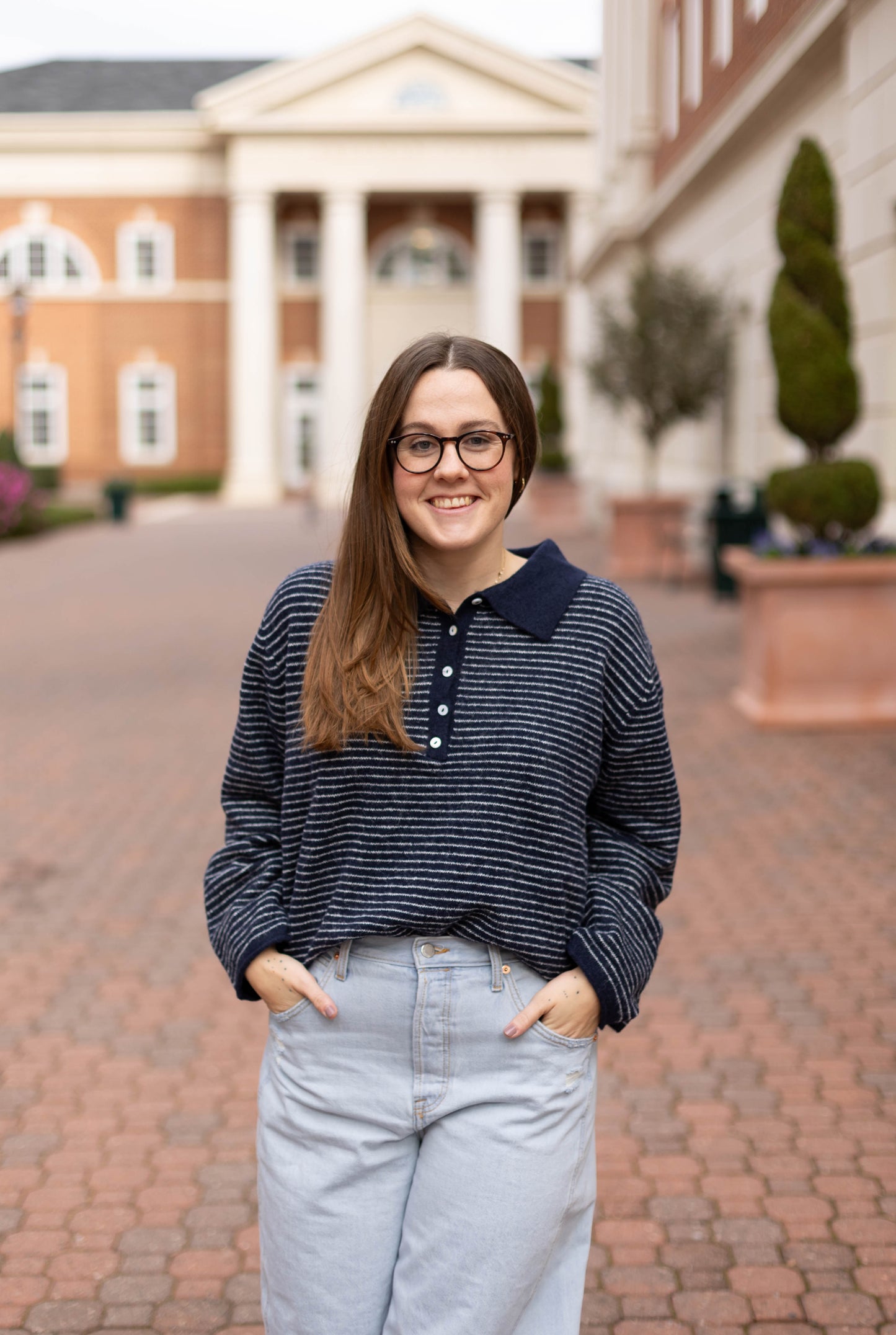 A woman with long brown hair and glasses stands outdoors on a brick walkway, wearing the Dogwood Cloth Anna Collared Henley and light jeans, with buildings and columns in the background.