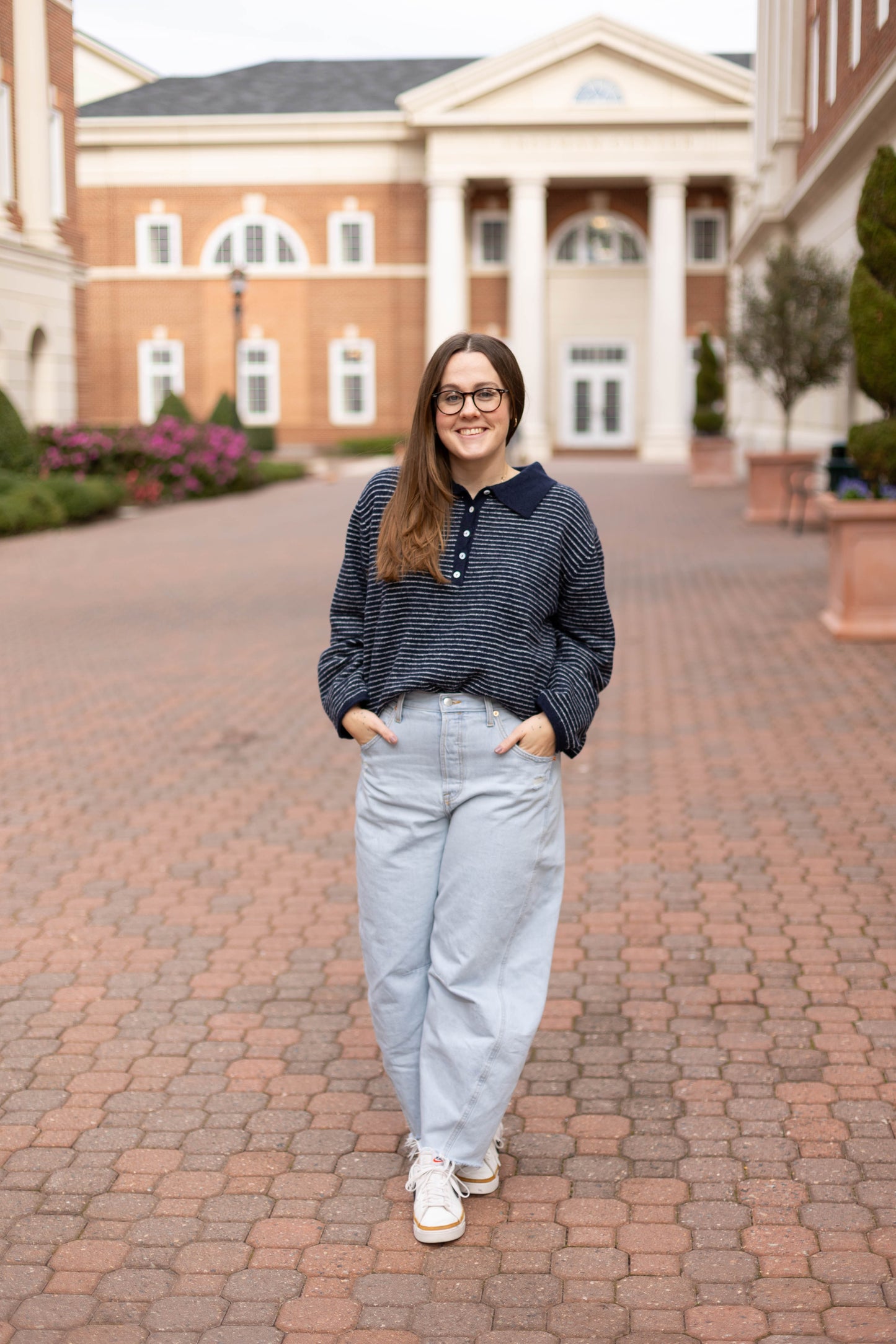 A woman with long brown hair and glasses wears a navy-and-white stripe Anna Collared Henley by Dogwood Cloth, standing on a brick walkway in front of a large brick building with white columns.