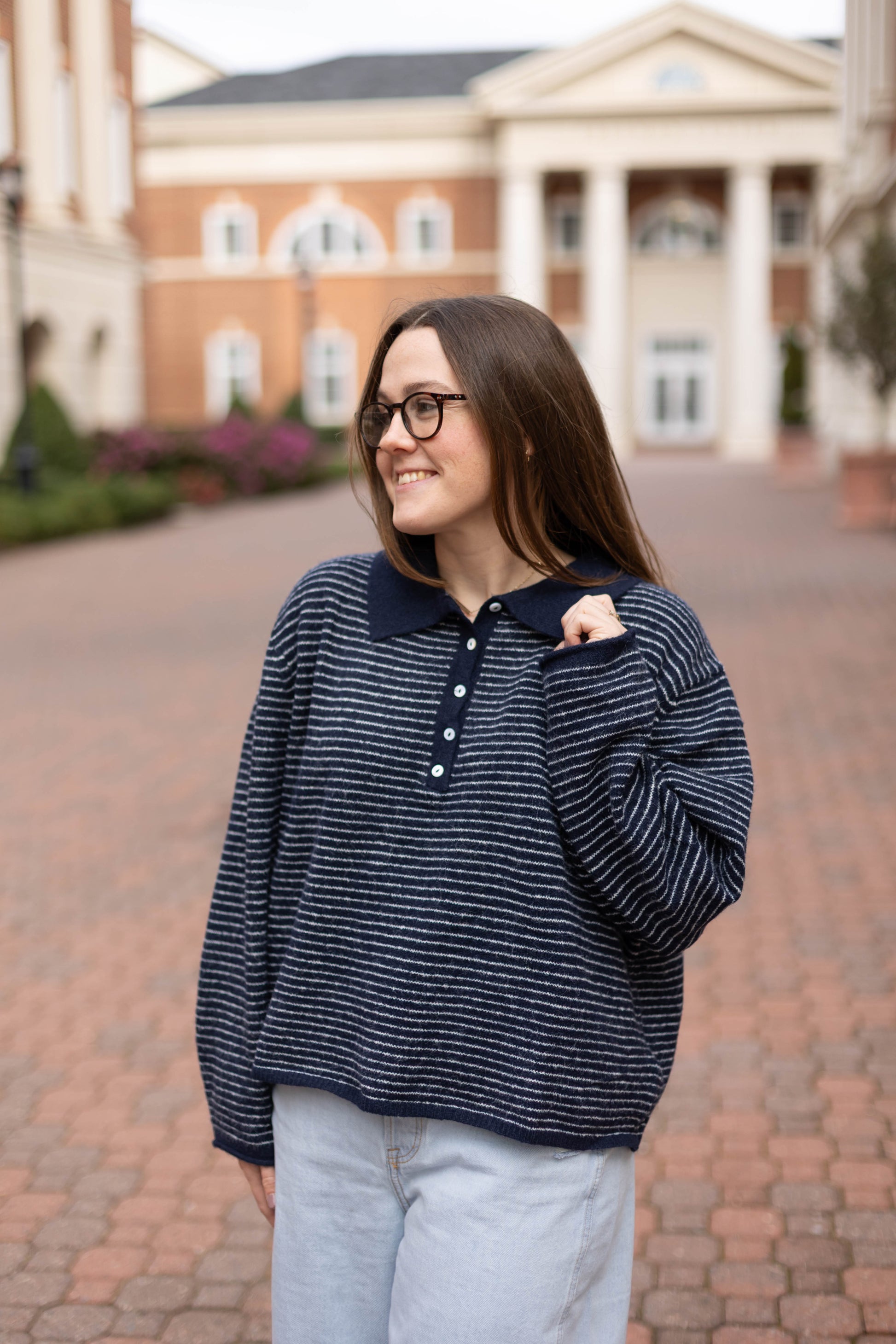 A woman in glasses and light jeans stands outdoors on a brick walkway, smiling, wearing the Dogwood Cloth Anna Collared Henley. A building is visible in the background.