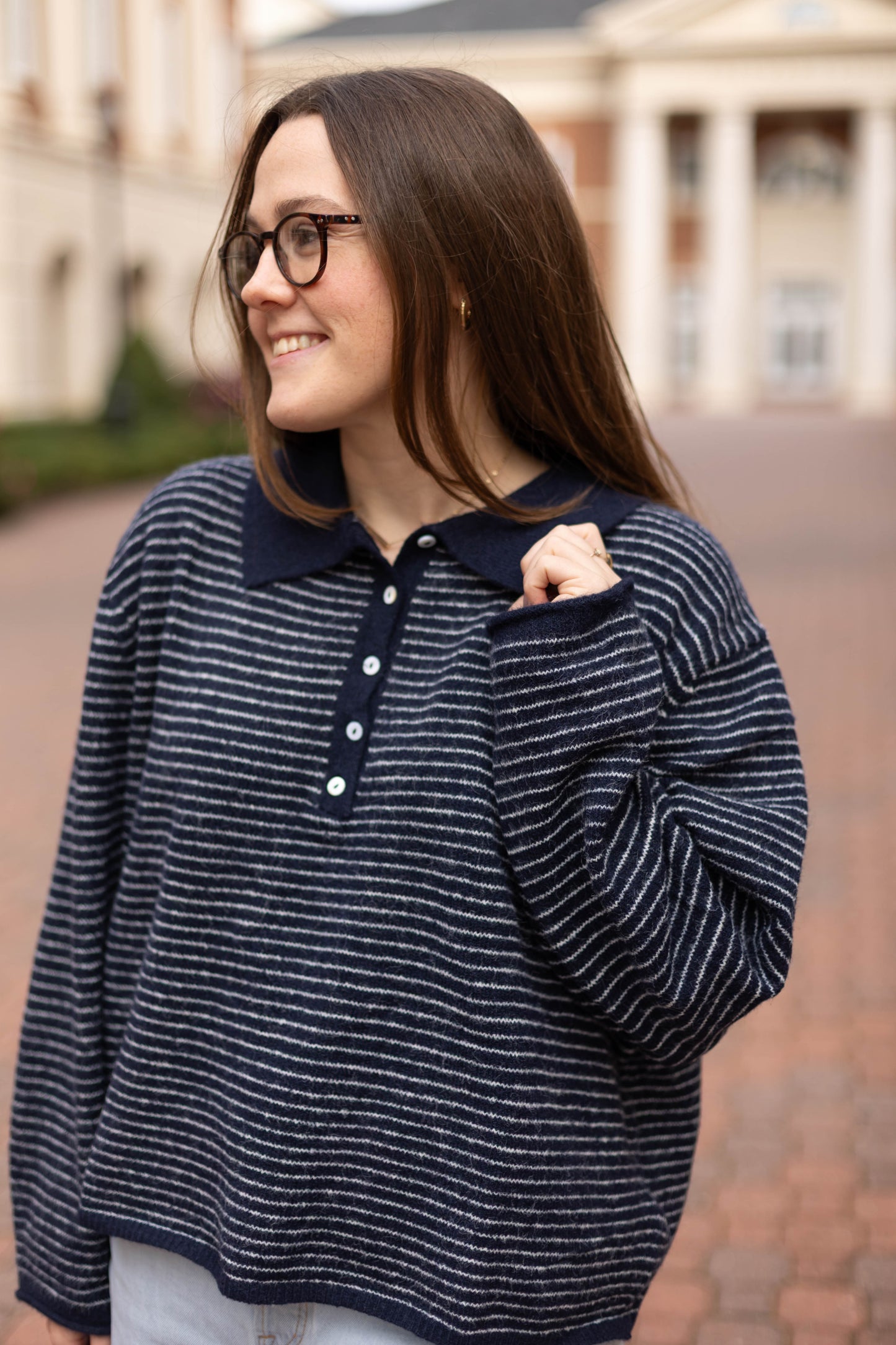 A woman wearing glasses and the Dogwood Cloth Anna Collared Henley, a navy-and-white stripe knit top, stands smiling outdoors on a brick walkway with a building featuring columns in the background.