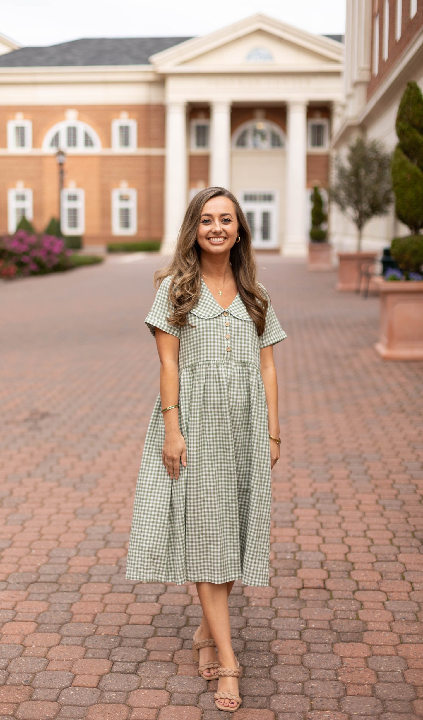 A woman smiles on a brick walkway in front of a grand building, wearing the Caroline Gingham Dress by Dogwood Cloth—a green dress featuring a Peter Pan collar.