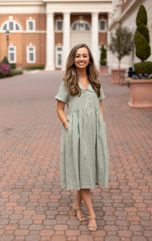 A woman smiles on a brick pathway in front of a grand building, wearing the Caroline Gingham Dress by Dogwood Cloth—a green dress with a Peter Pan collar.