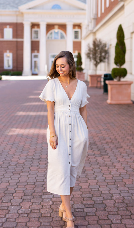 A woman in the Chelsea Linen Midi by Dogwood Cloth stands on a brick walkway, smiling as she looks down, with a building and trees in the background.