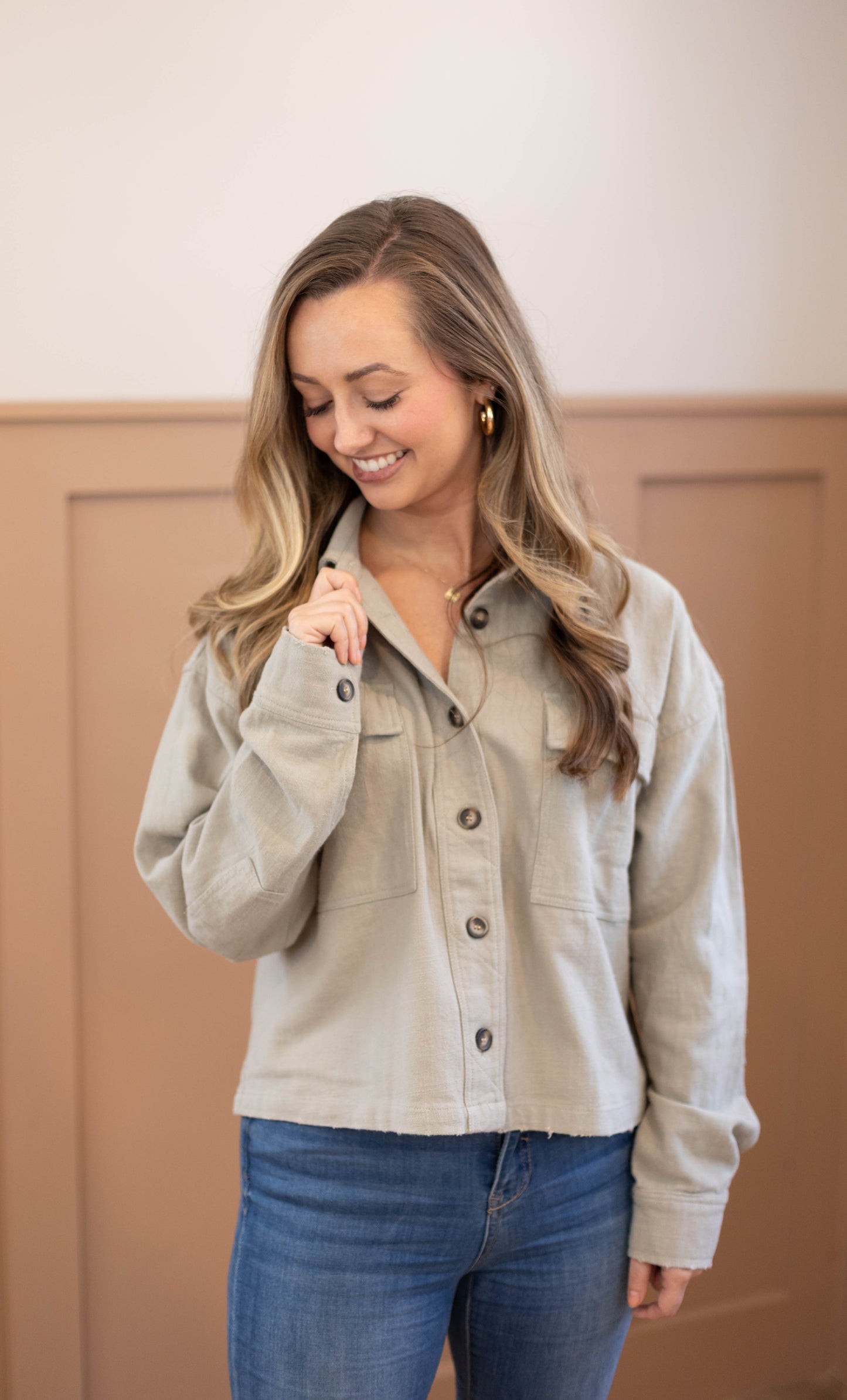 A woman with long, wavy hair stands indoors, smiling and looking down while wearing the Ruth Cropped Jacket by Dogwood Cloth in light gray, paired with blue jeans.