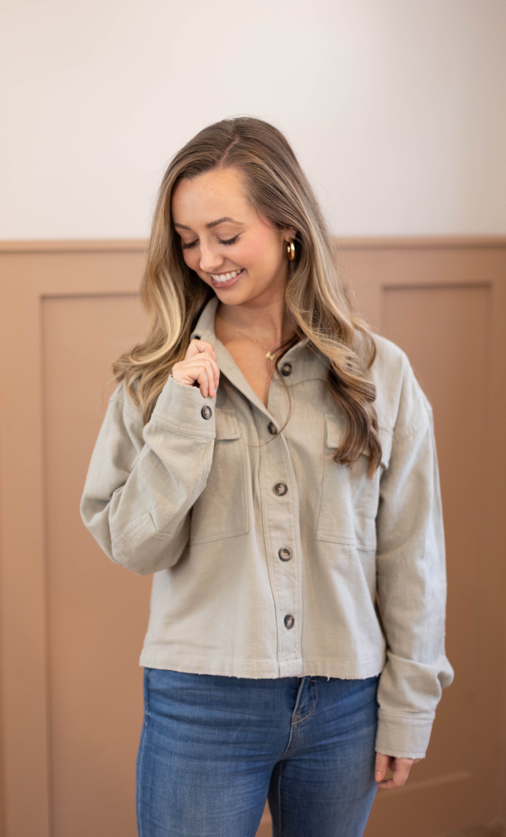 A woman with long, wavy hair stands indoors, smiling and looking down while wearing the Ruth Cropped Jacket by Dogwood Cloth in light gray, paired with blue jeans.