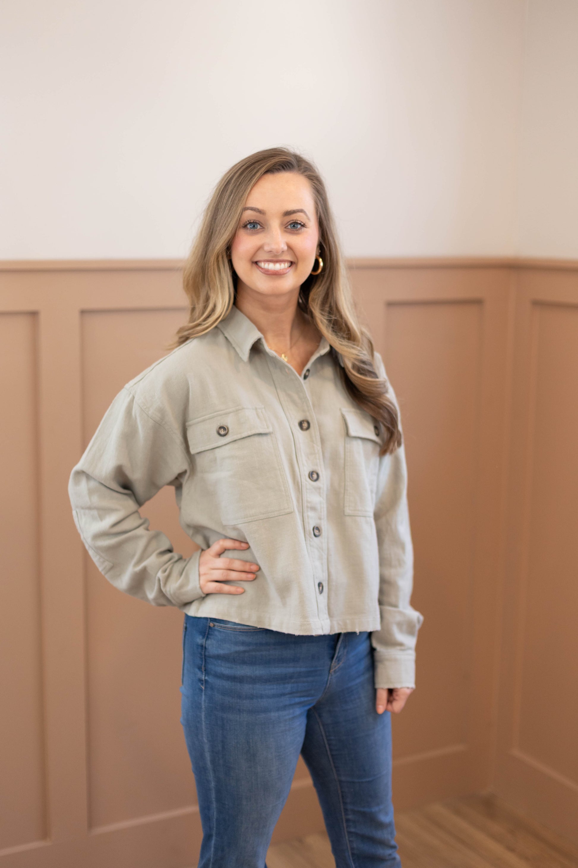 A woman smiles indoors with one hand on her hip, wearing blue jeans and the Dogwood Cloth Ruth Cropped Jacket in light gray (100% cotton) in front of a beige wainscoted wall.