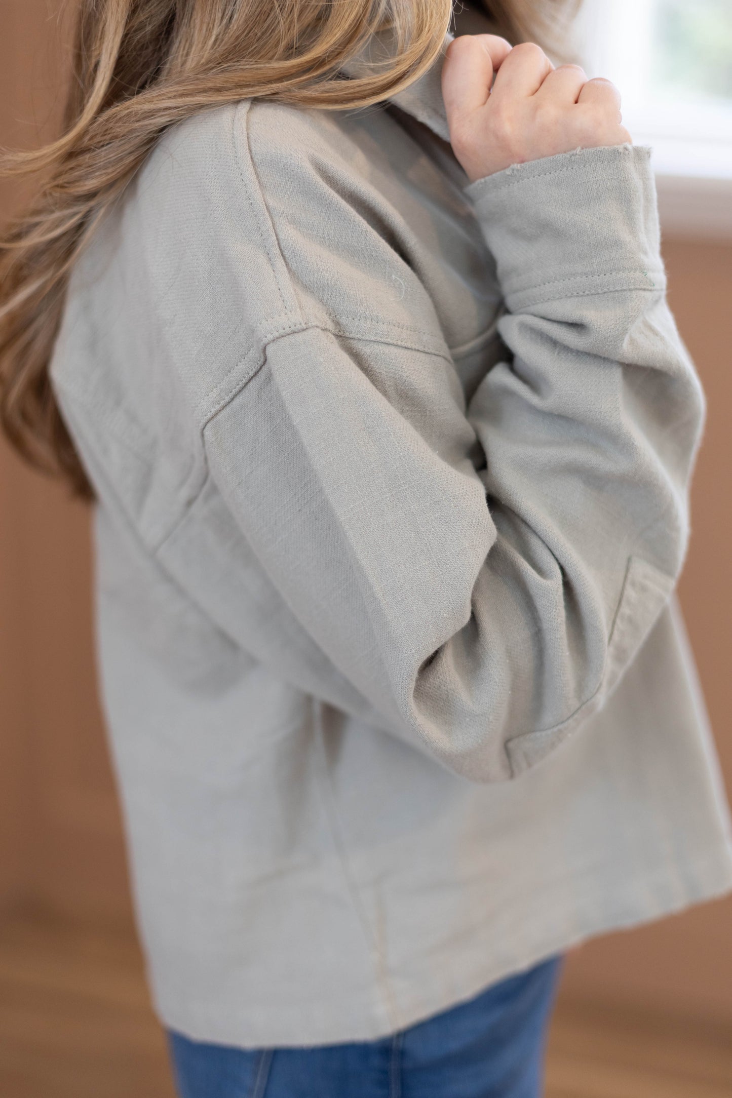 A person with long hair wears the Dogwood Cloth Ruth Cropped Jacket in light gray and blue jeans, holding the collar of the 100% cotton women's jacket with one hand.