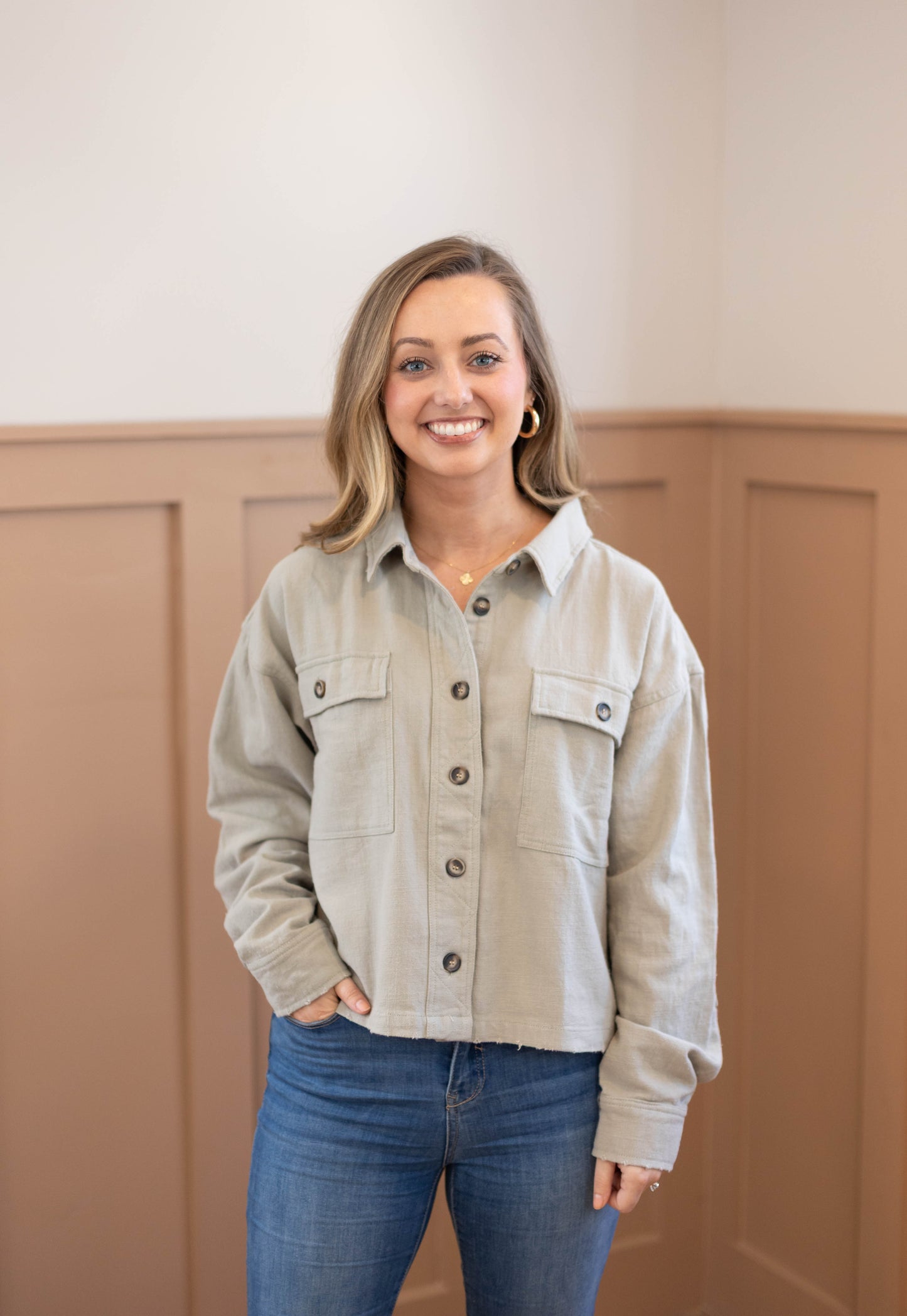 A woman with shoulder-length blonde hair, wearing the Ruth Cropped Jacket by Dogwood Cloth over a light shirt and blue jeans, stands indoors against tan paneled walls, smiling at the camera.
