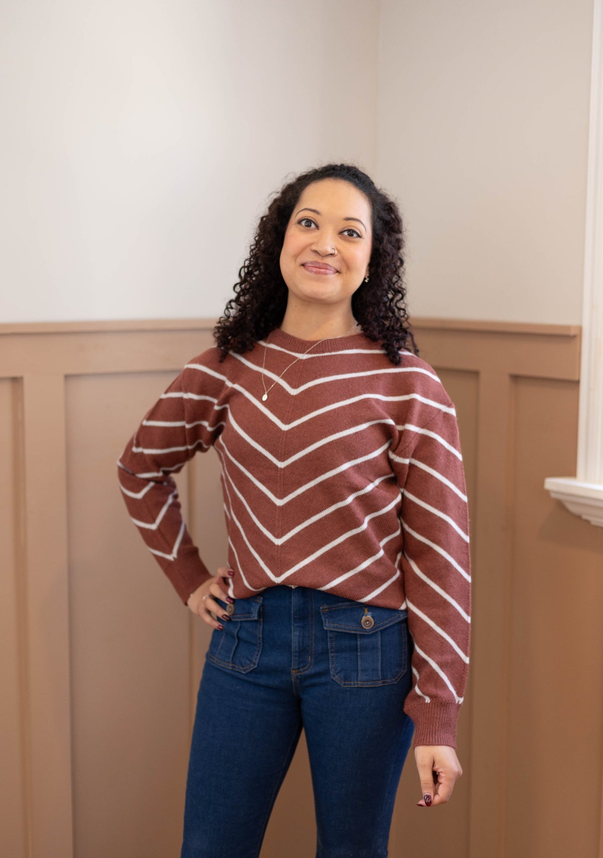 A woman with curly hair stands indoors wearing the Dogwood Cloth Hailey Sweater with brown stripes and jeans, one hand on her hip and a neutral expression.