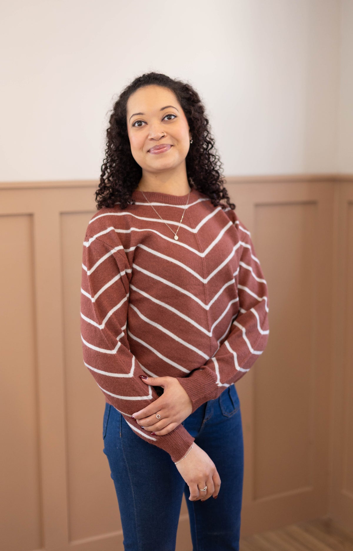 A woman with curly hair wears the Dogwood Cloth Hailey Sweater, a brown and white striped chevron design, paired with blue jeans as she stands indoors, smiling gently with her hands folded.