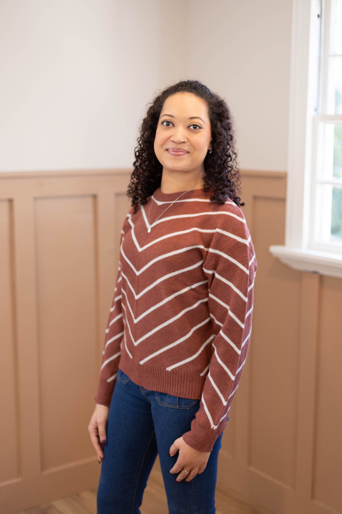 A woman with curly hair wears the Dogwood Cloth Hailey Sweater in brown and white stripes with blue jeans, standing indoors by a window and looking at the camera.