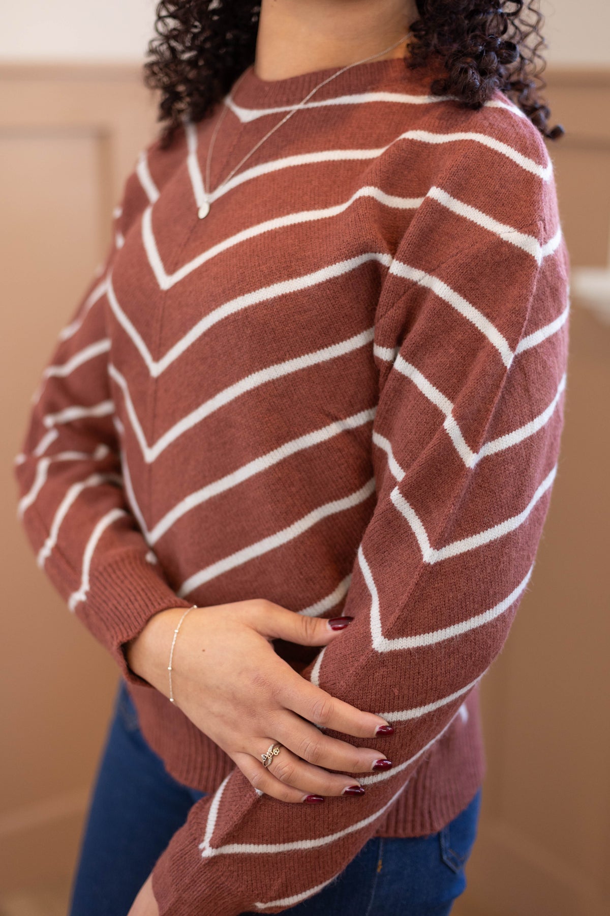 A person wearing the Dogwood Cloth Hailey Sweater in rust with white stripes, paired with blue jeans and jewelry, stands with one arm crossed over the other.