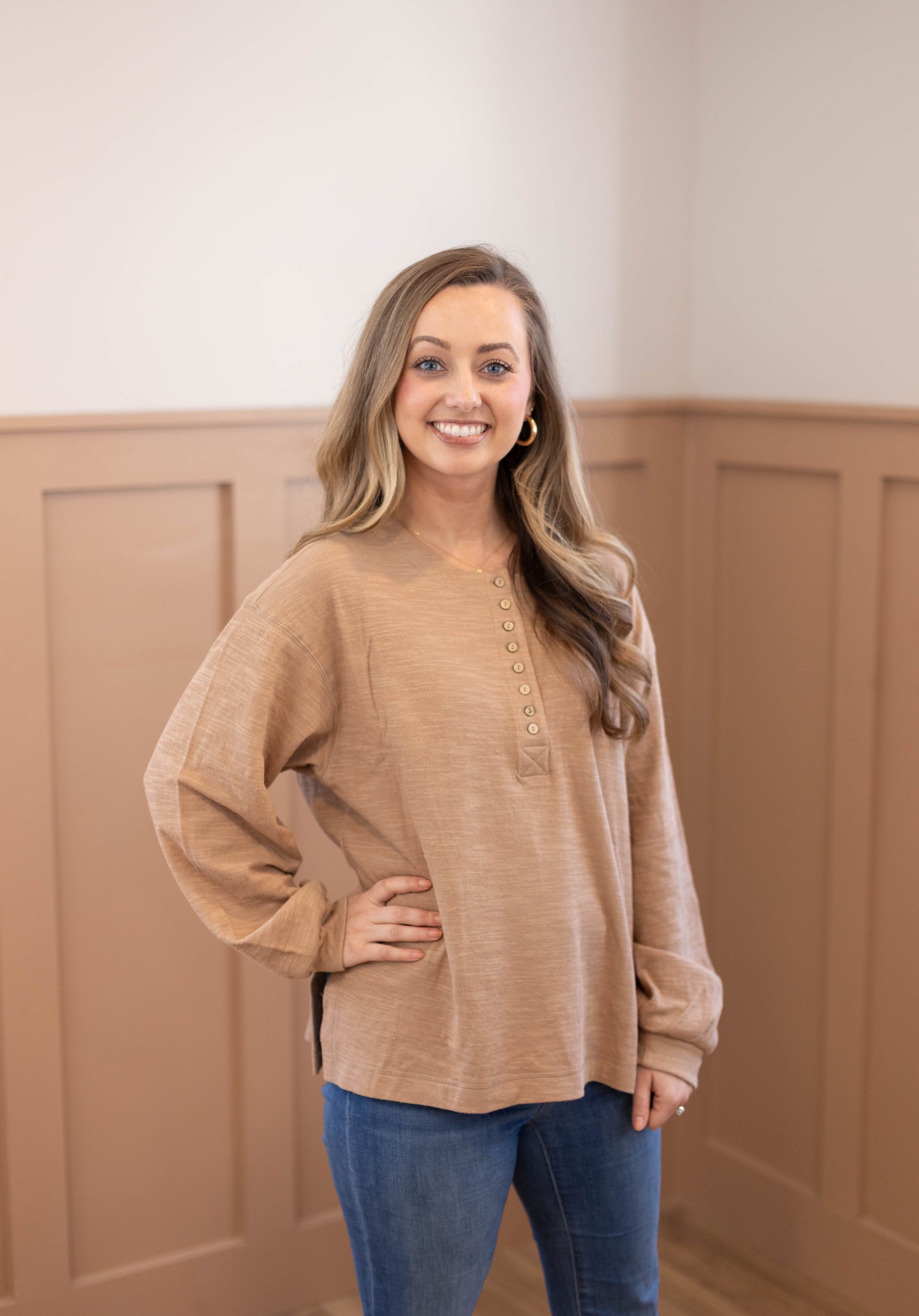 A woman with long hair, wearing the Dogwood Cloth Clark Everyday Henley in tan and blue jeans, stands indoors before tan wainscoting, smiling at the camera.
