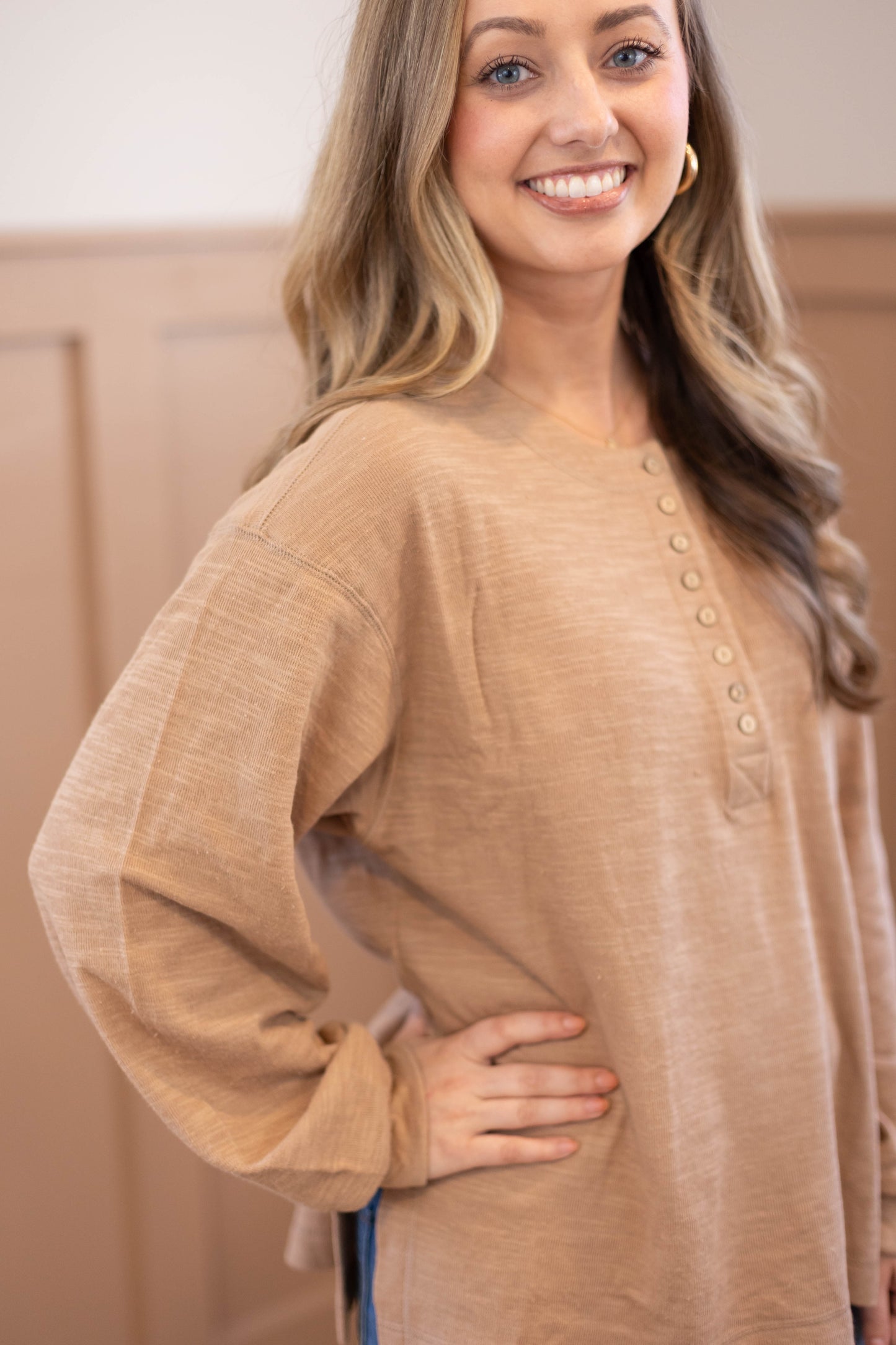 A woman with long, wavy hair smiles at the camera, wearing the Dogwood Cloth Clark Everyday Henley in tan—a long sleeve, nursing-friendly top—with her hand on her hip.
