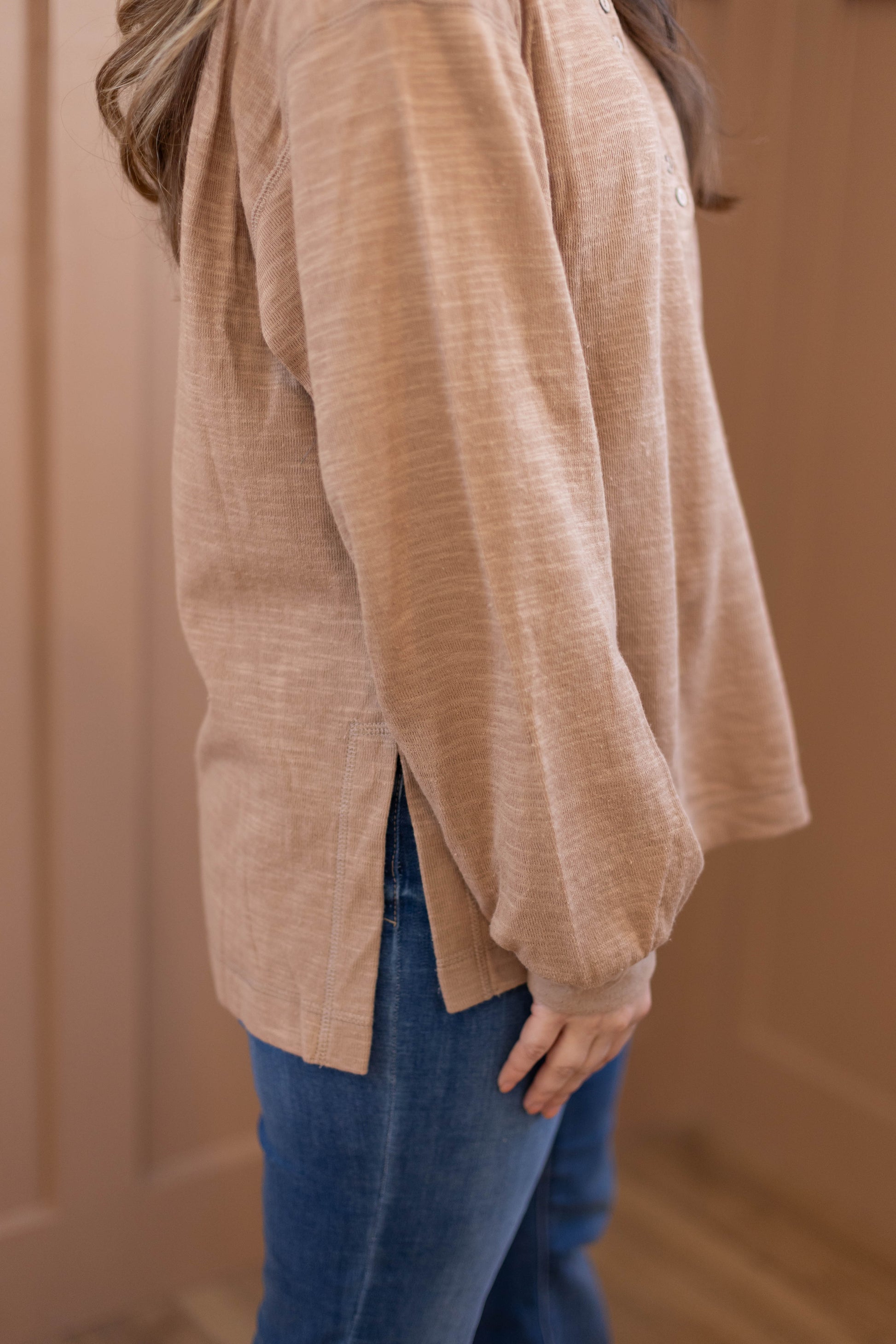 A person stands indoors by a light brown wall wearing Dogwood Cloth's Clark Everyday Henley—a lightweight, long-sleeved cotton top with side slits—paired with blue jeans.