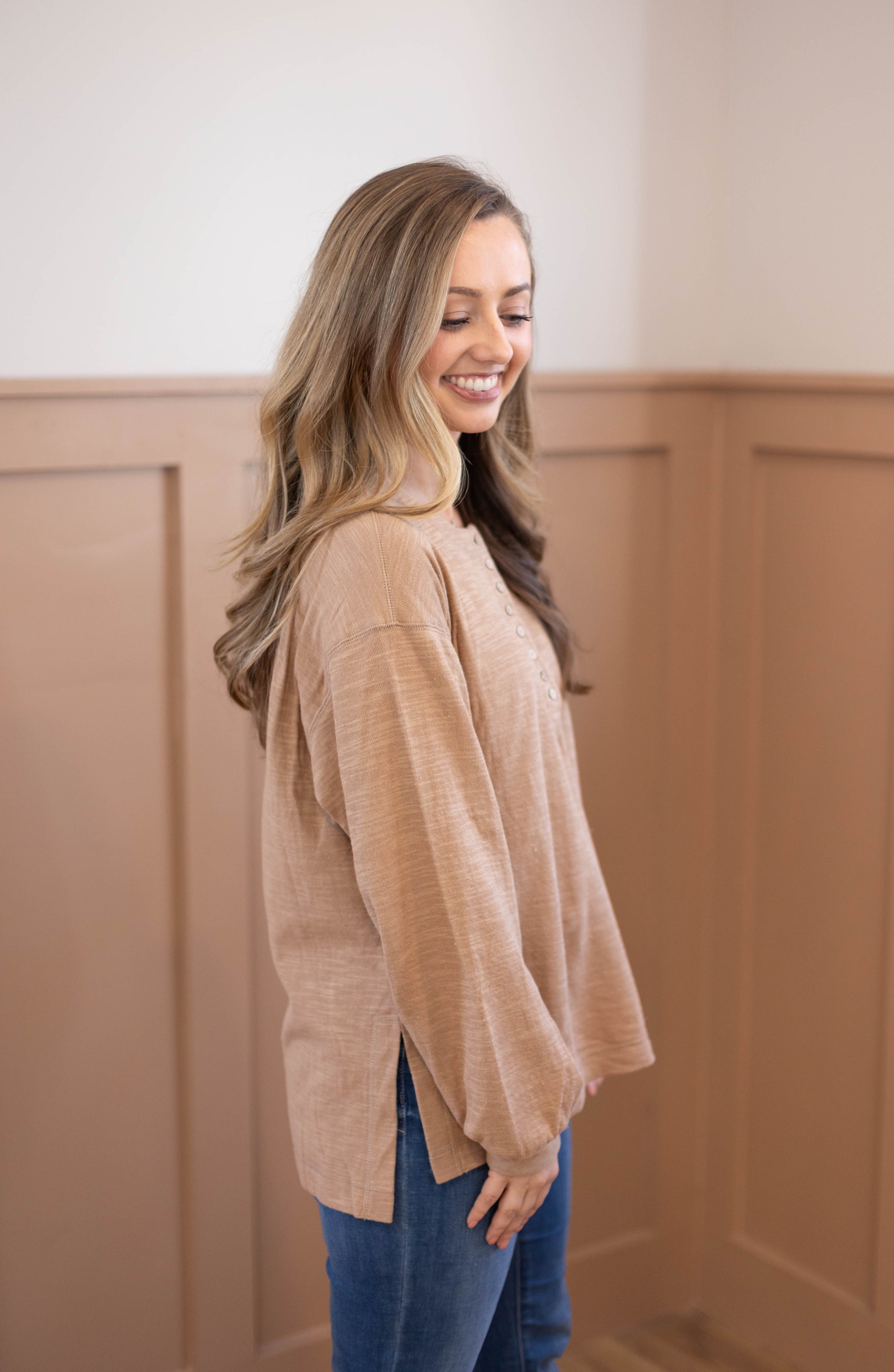 A woman stands sideways, smiling, wearing the Dogwood Cloth Clark Everyday Henley—a nursing-friendly, lightweight cotton top—with blue jeans in a beige-paneled room.
