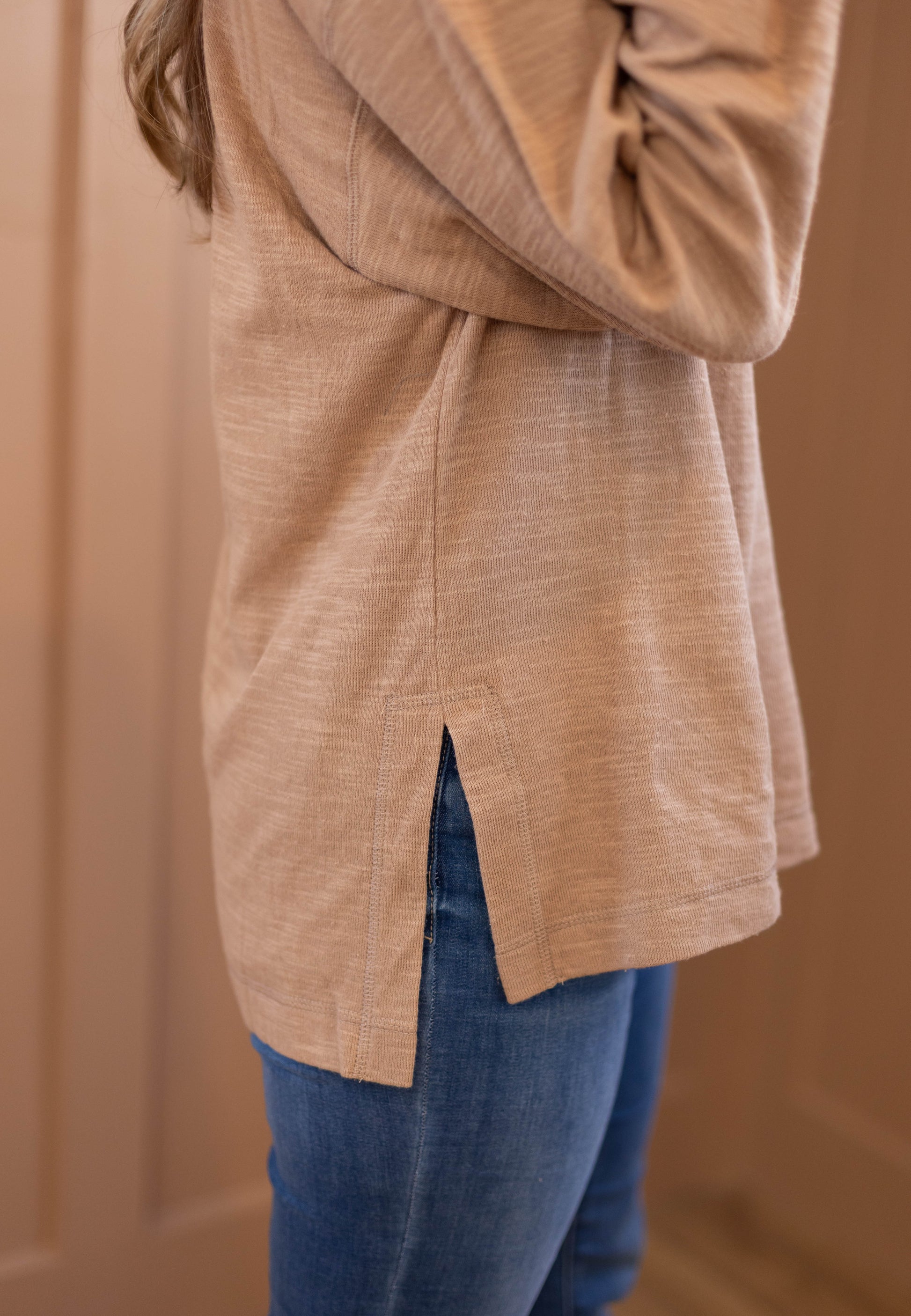 A person in the Dogwood Cloth Clark Everyday Henley, a long sleeve top with a side slit, pairs it with blue jeans while standing sideways against a light brown background—an effortless style, ideal for nursing moms.